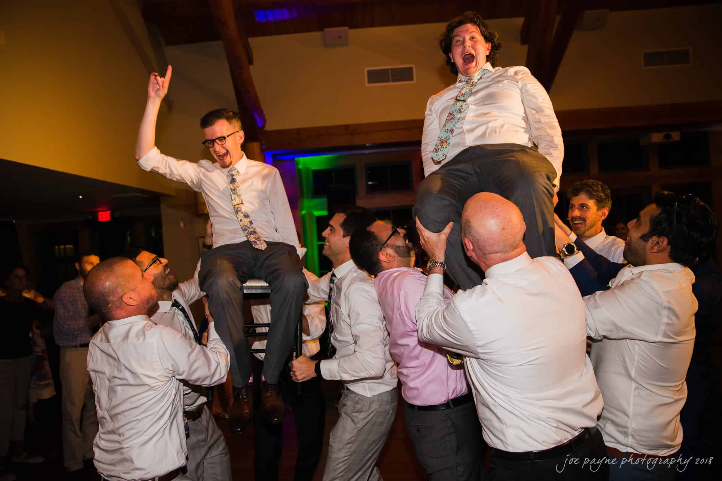 A group of people celebrating at a party. Two men are being lifted on chairs by friends. They are laughing and appear to be at a joyful event, possibly a wedding reception. The room is warmly lit, creating a festive atmosphere.