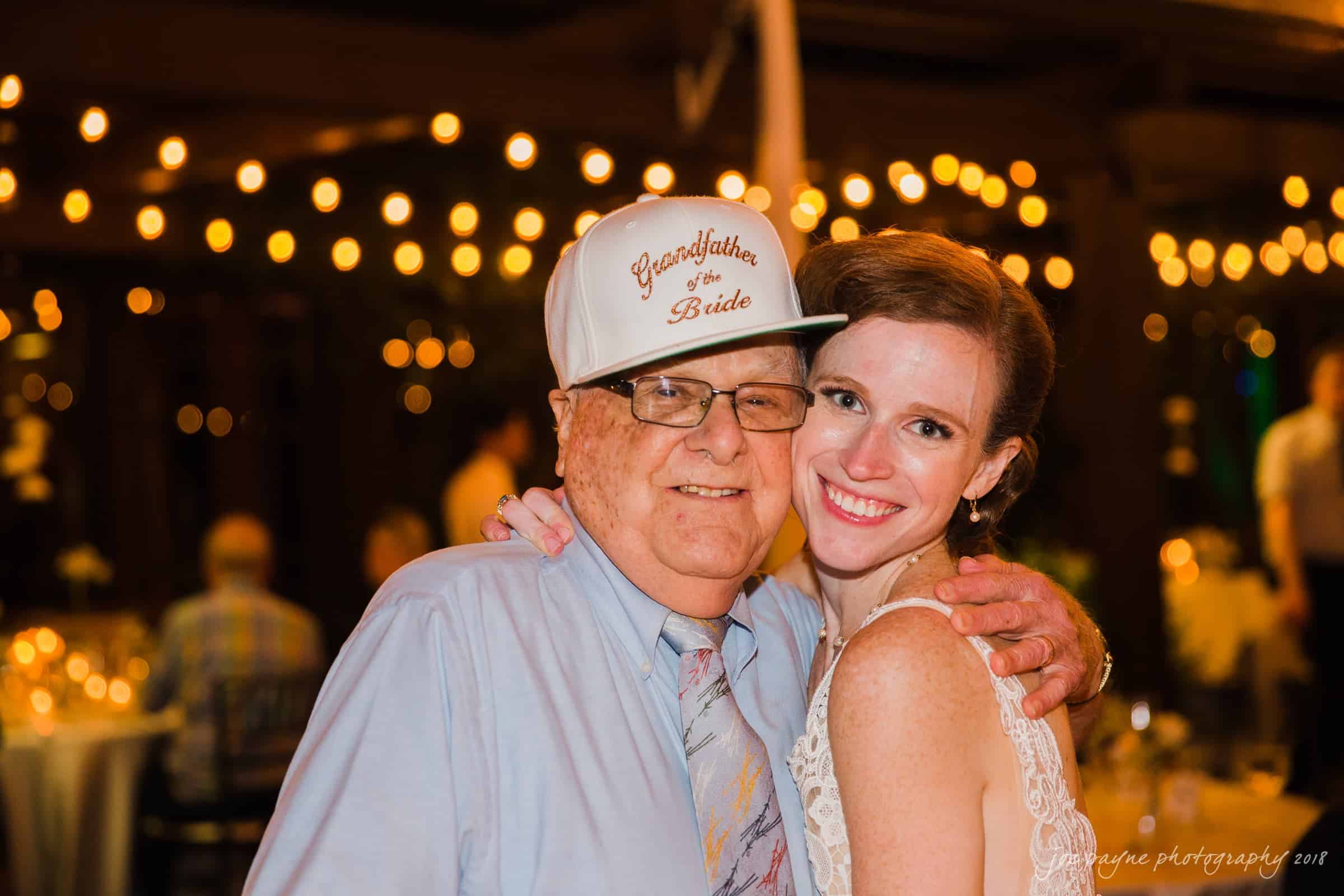 A smiling bride in a white dress poses with an older man wearing glasses, a tie, and a cap that reads Grandfather of the Bride, against a backdrop of soft, glowing string lights at a festive event.