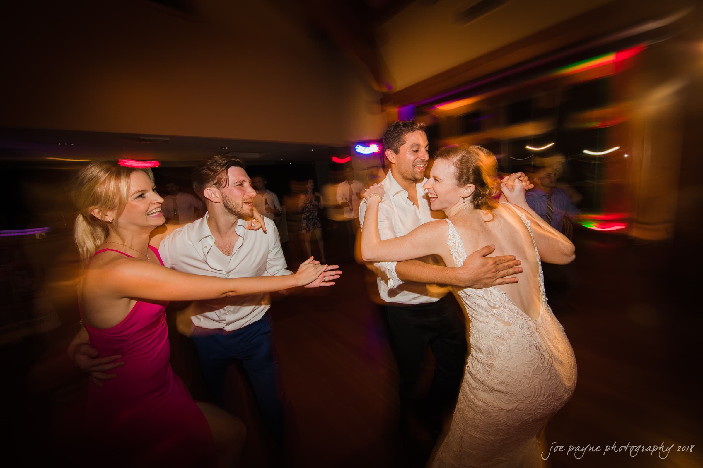 A joyful group of four people, including a couple in wedding attire, dance energetically on a dimly lit dance floor. The ambient lighting creates a festive and lively atmosphere.