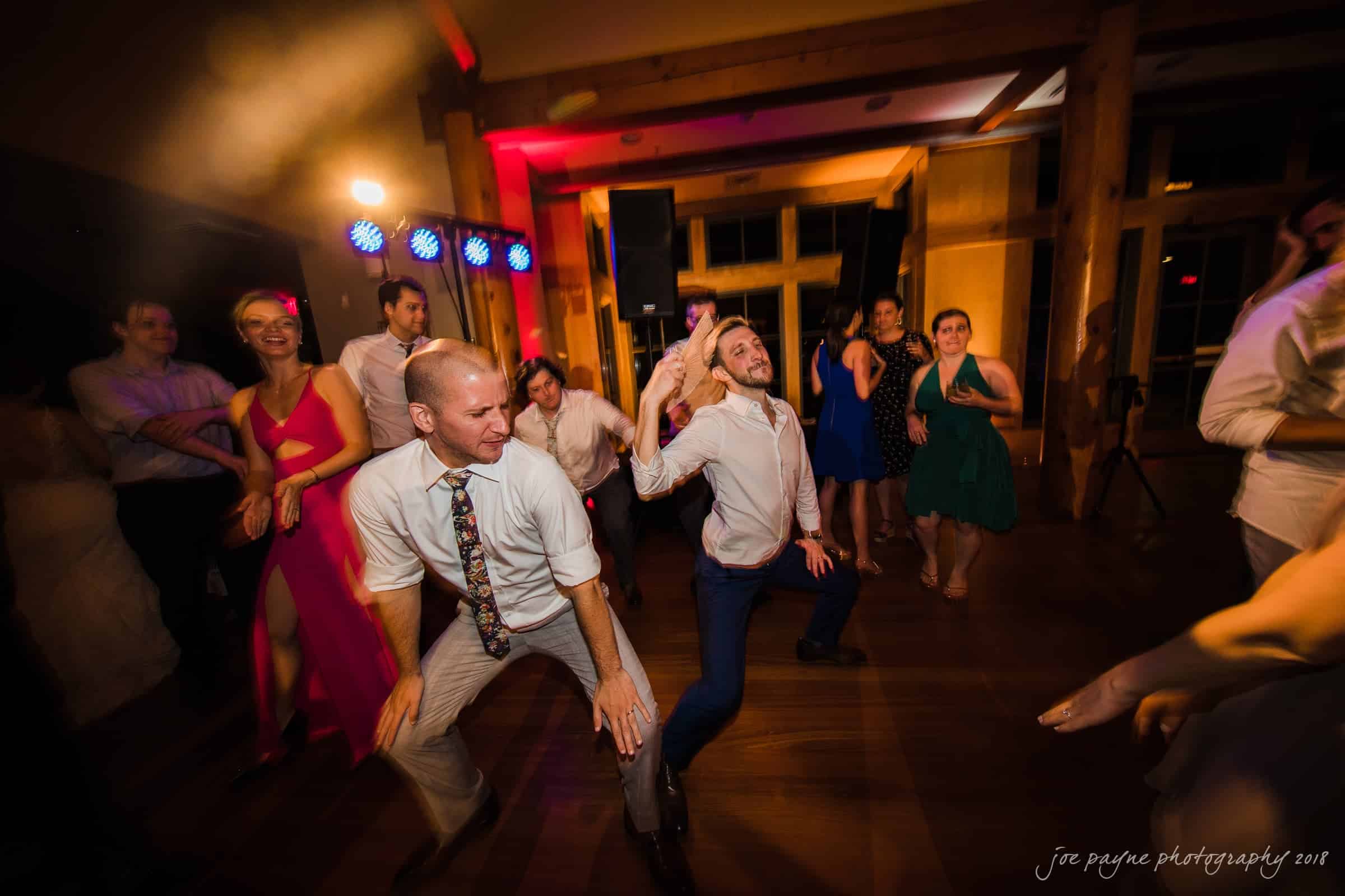 A lively group of people dancing energetically at a party. The room is warmly lit, creating a cheerful and celebratory atmosphere. Two men in the foreground dance enthusiastically, surrounded by others enjoying the moment.