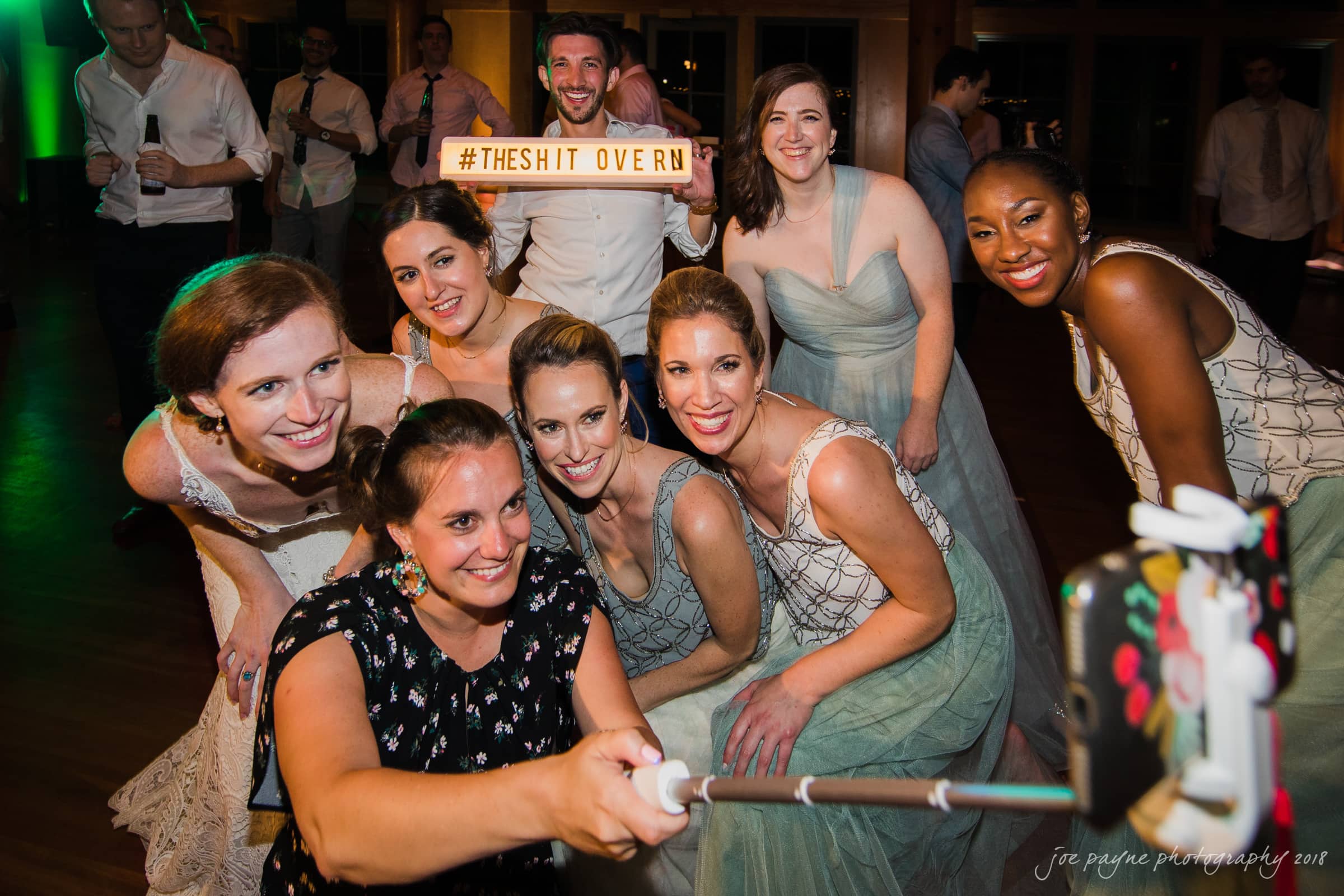 A group of eight people dressed in formal attire take a selfie at an event. They smile and pose, while one person holds a light-up sign reading #THESHITOVER. The background shows more guests in a dimly lit venue.