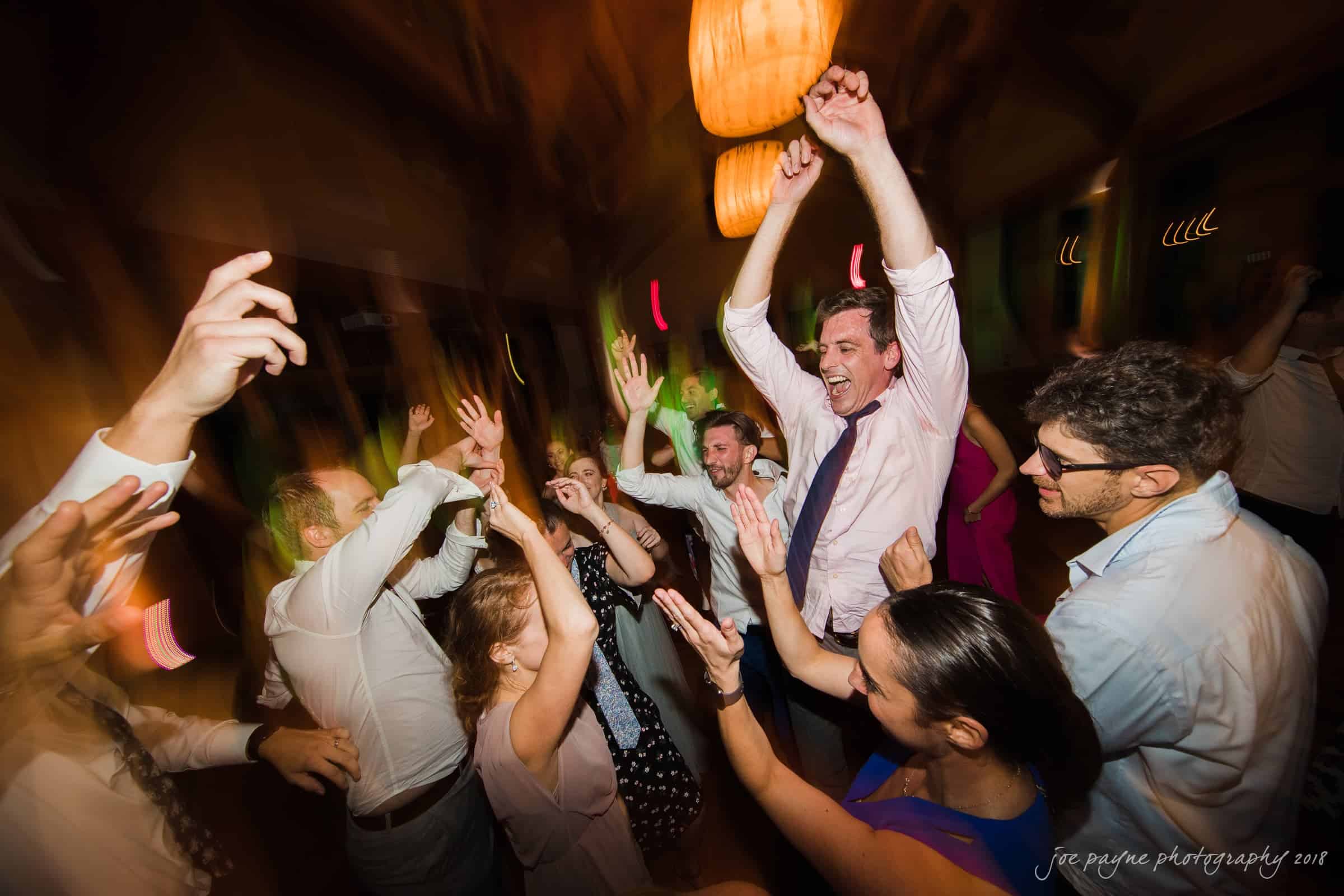 A group of people are joyfully dancing with their hands in the air at a lively celebration. The lighting creates a dynamic and energetic atmosphere.