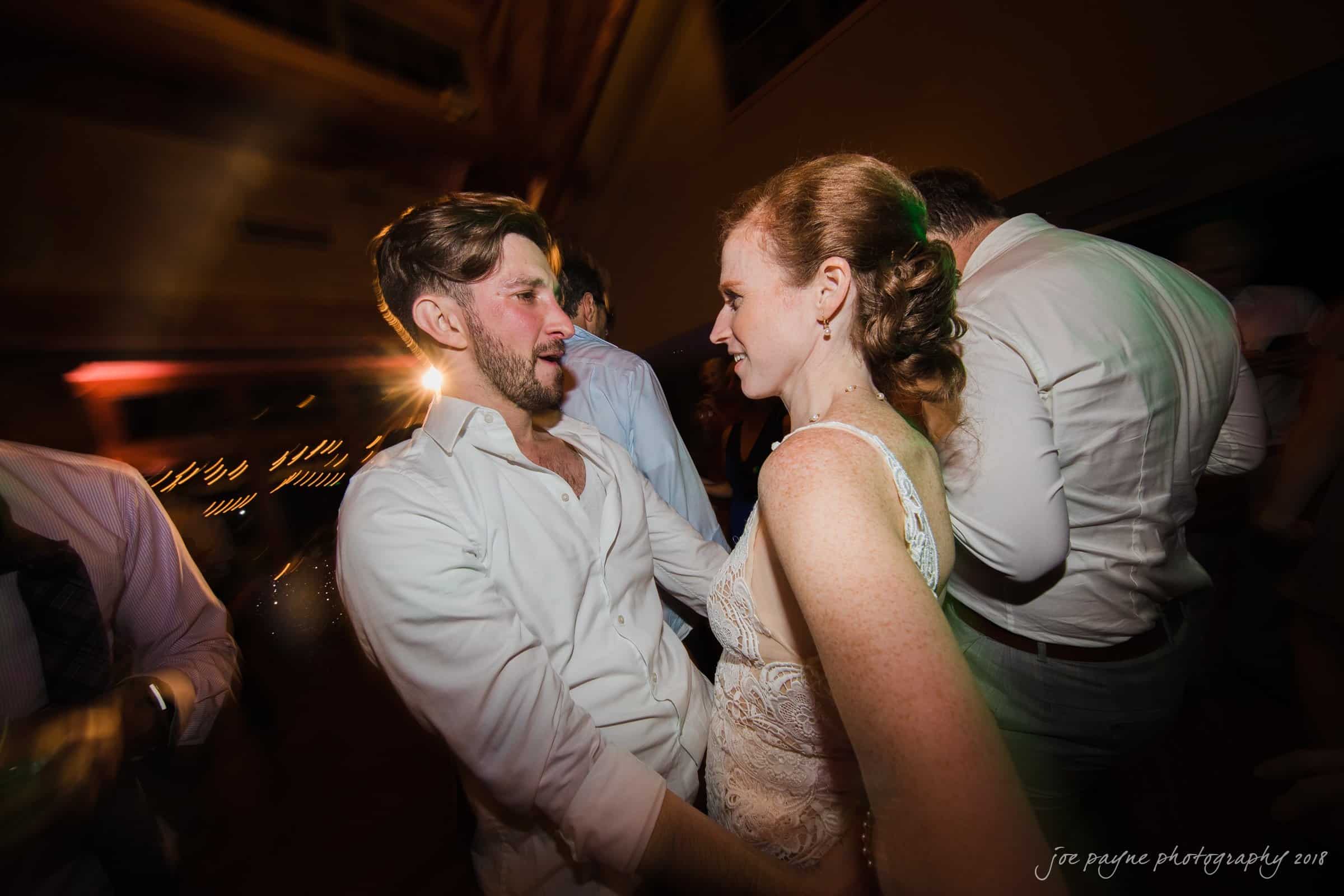 A couple is dancing closely at an indoor event. The man is wearing a white shirt, and the woman is in a lace dress. The background is softly lit, creating a warm and intimate atmosphere. Other guests are visible but blurred.