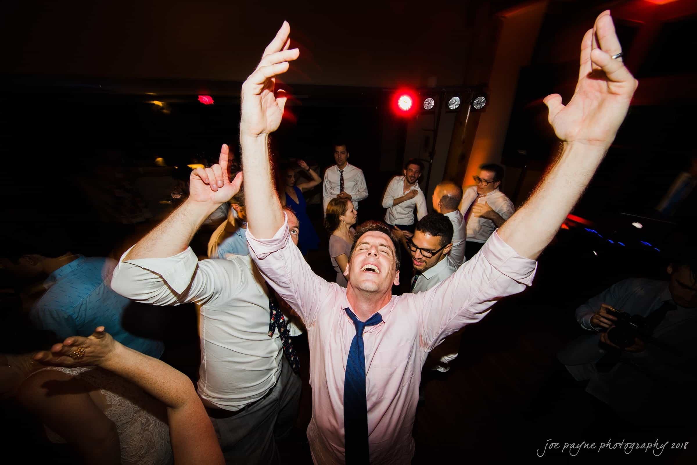 A group of people dancing enthusiastically at an indoor party. The central figure, a man in a pink shirt and tie, is raising his arms joyfully. The setting is dimly lit with colored lights creating a lively atmosphere.