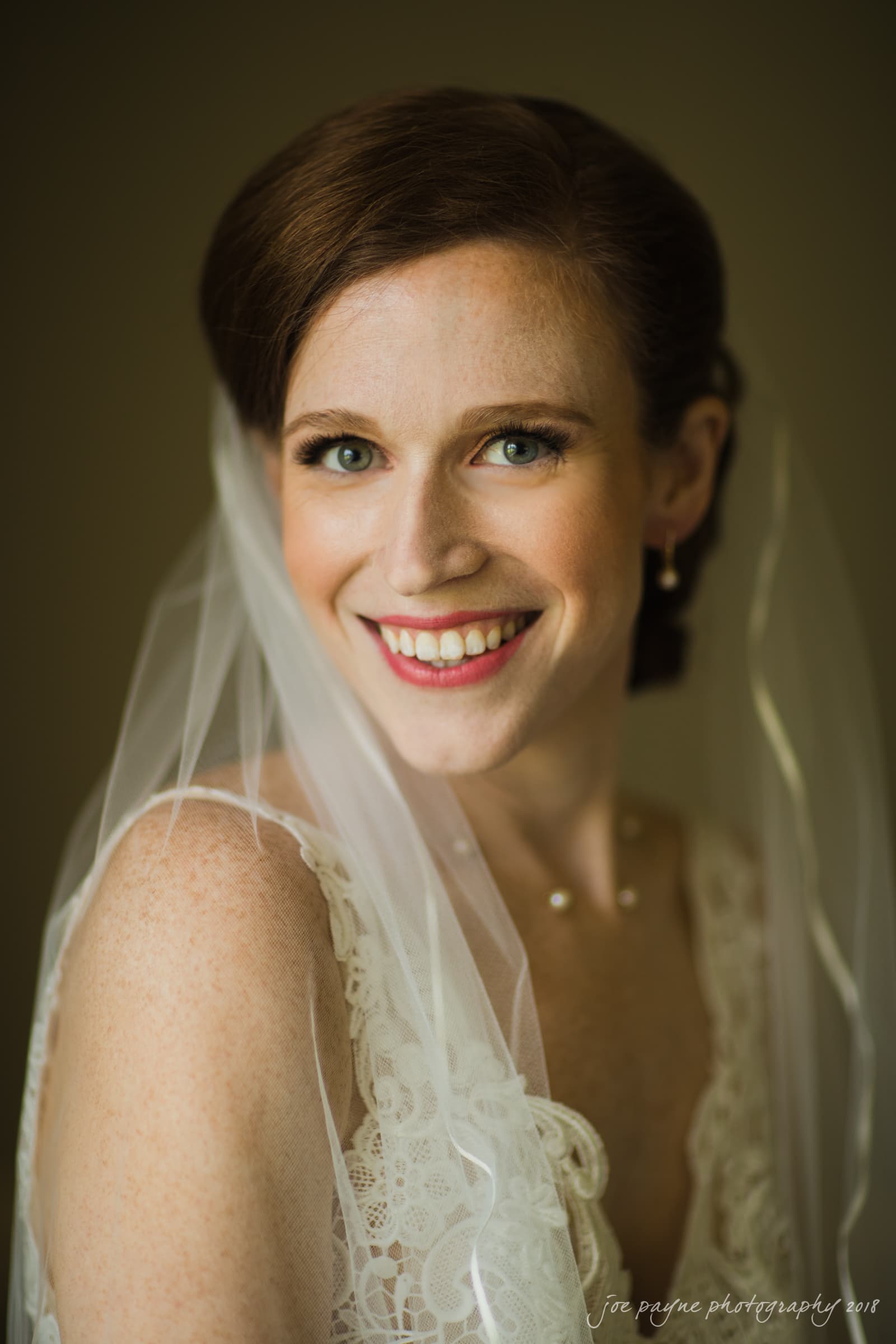 A smiling bride with red hair wears a lace wedding dress and a veil. The soft lighting highlights her freckles and elegant appearance.