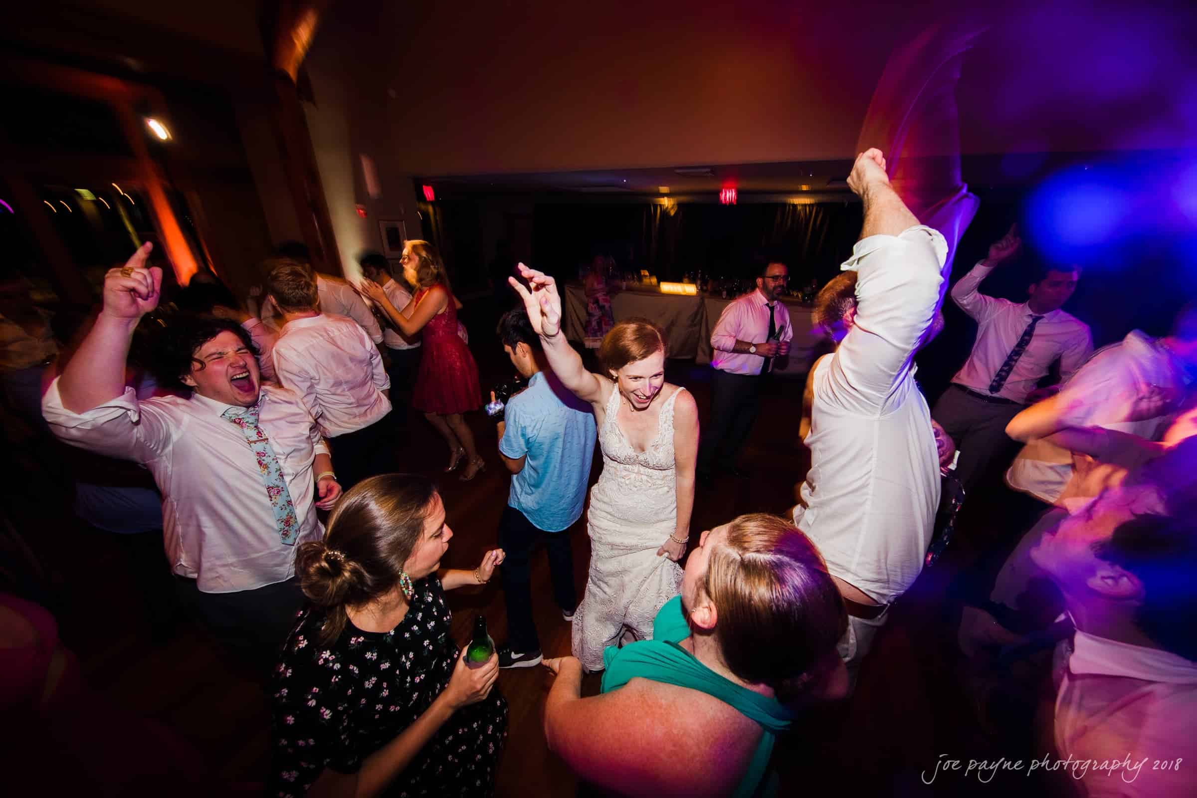 A lively wedding reception with people dancing joyfully on a dimly lit dance floor. The bride is at the center in a white gown, surrounded by guests celebrating with raised hands and smiles. Colorful lights enhance the festive atmosphere.