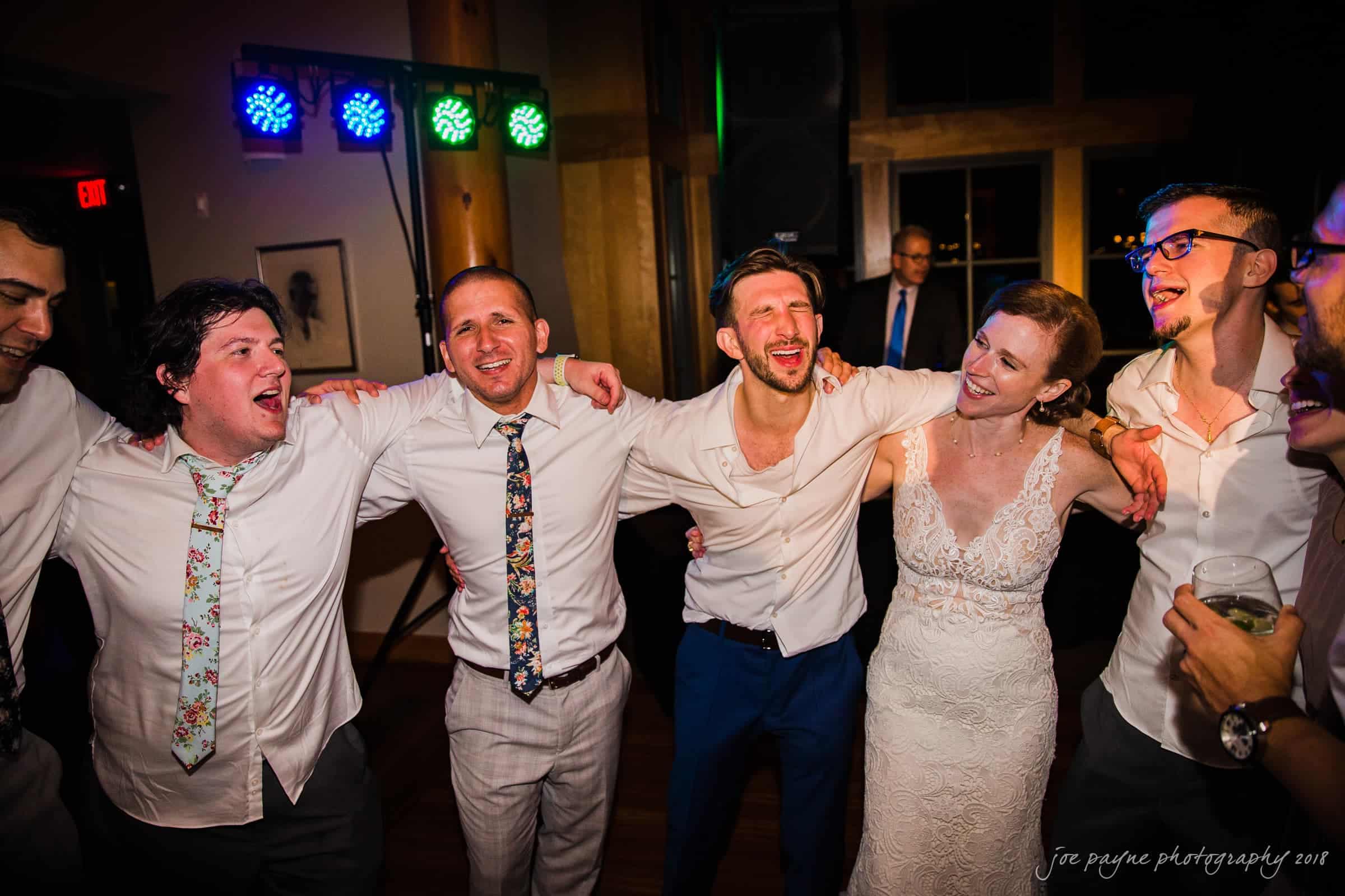 A group of people, including a woman in a wedding dress, are joyfully embracing and singing together at a celebration. The lively atmosphere is enhanced by colorful lights in the background.