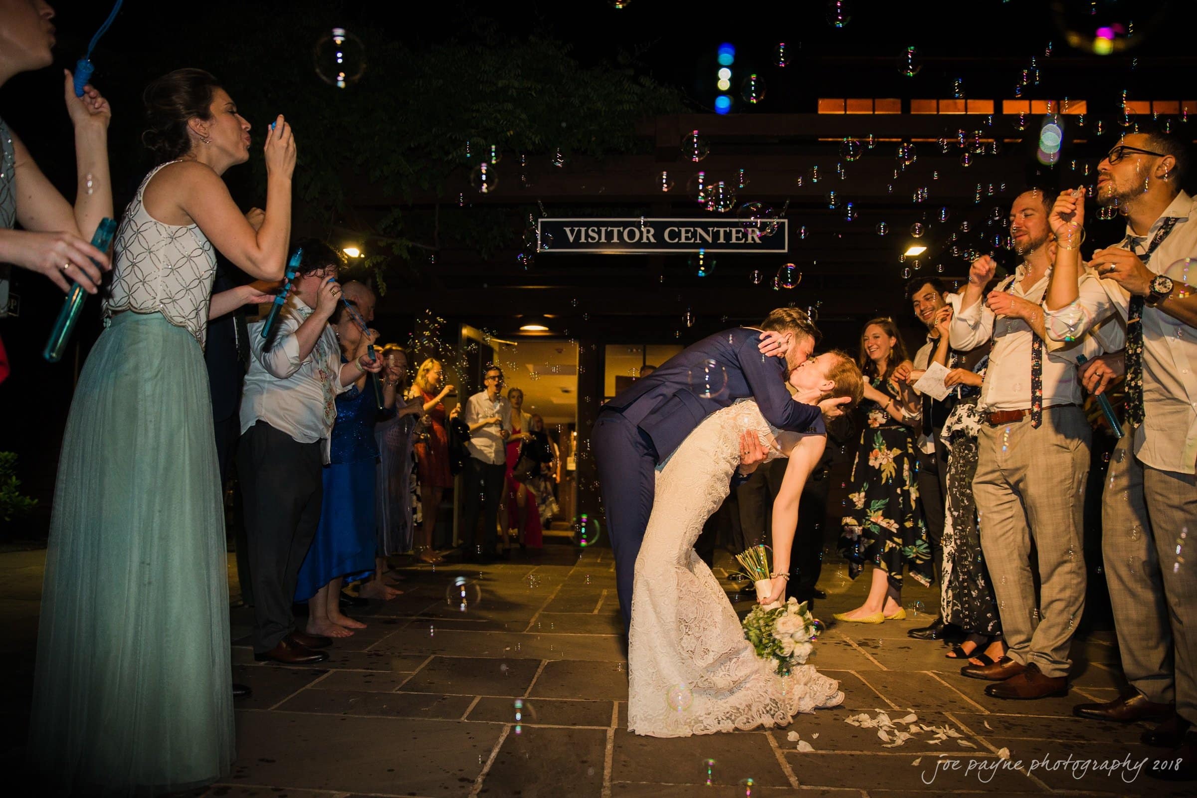 A newlywed couple shares a romantic kiss under a shower of bubbles at night. They are surrounded by applauding and cheering guests, forming a joyful path outside a visitor center. The scene is festive and filled with celebration.