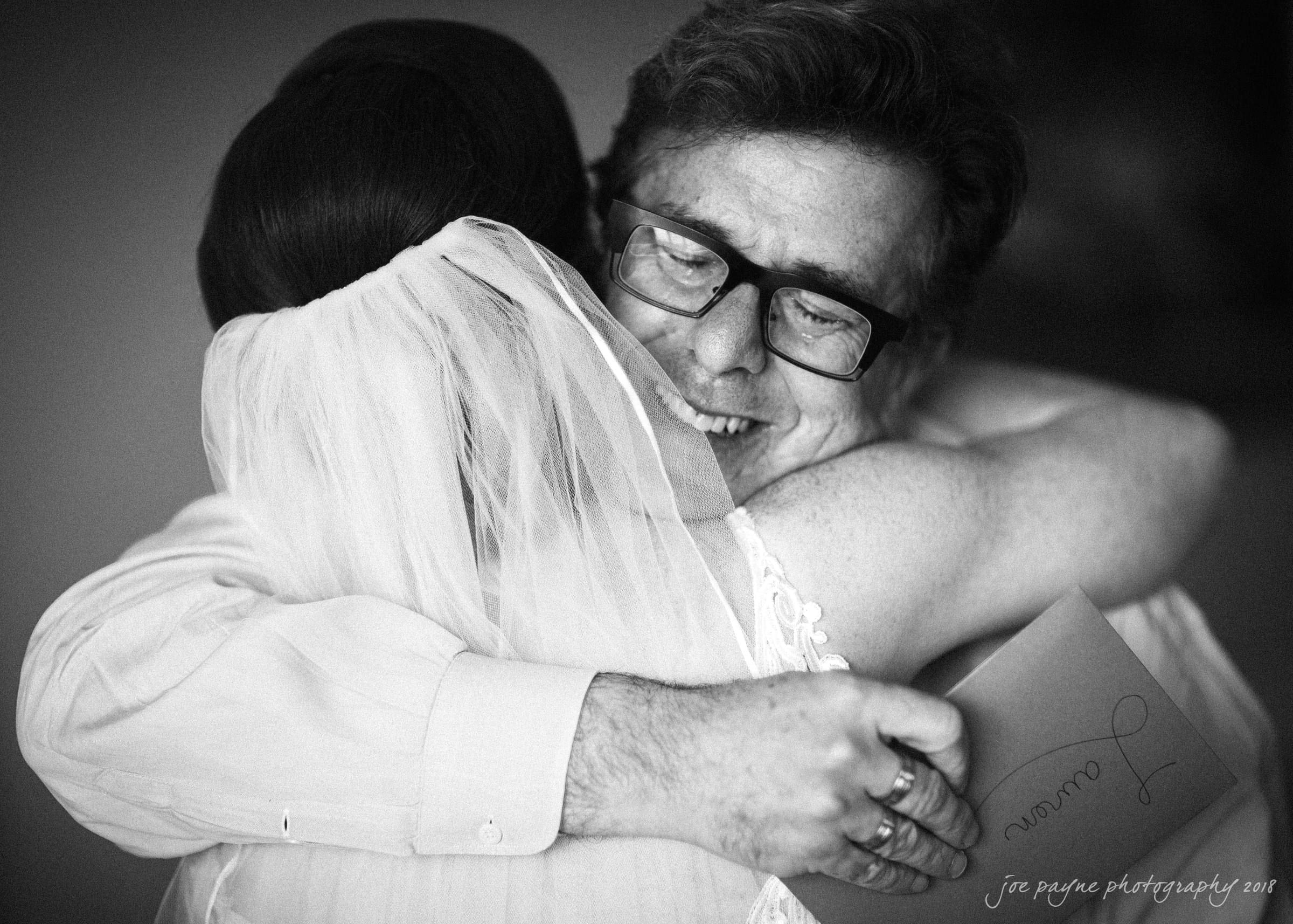 A bride and a man share a warm embrace, both smiling with closed eyes. The bride is wearing a veil, and the man is holding a card. The image captures a joyful and touching moment.