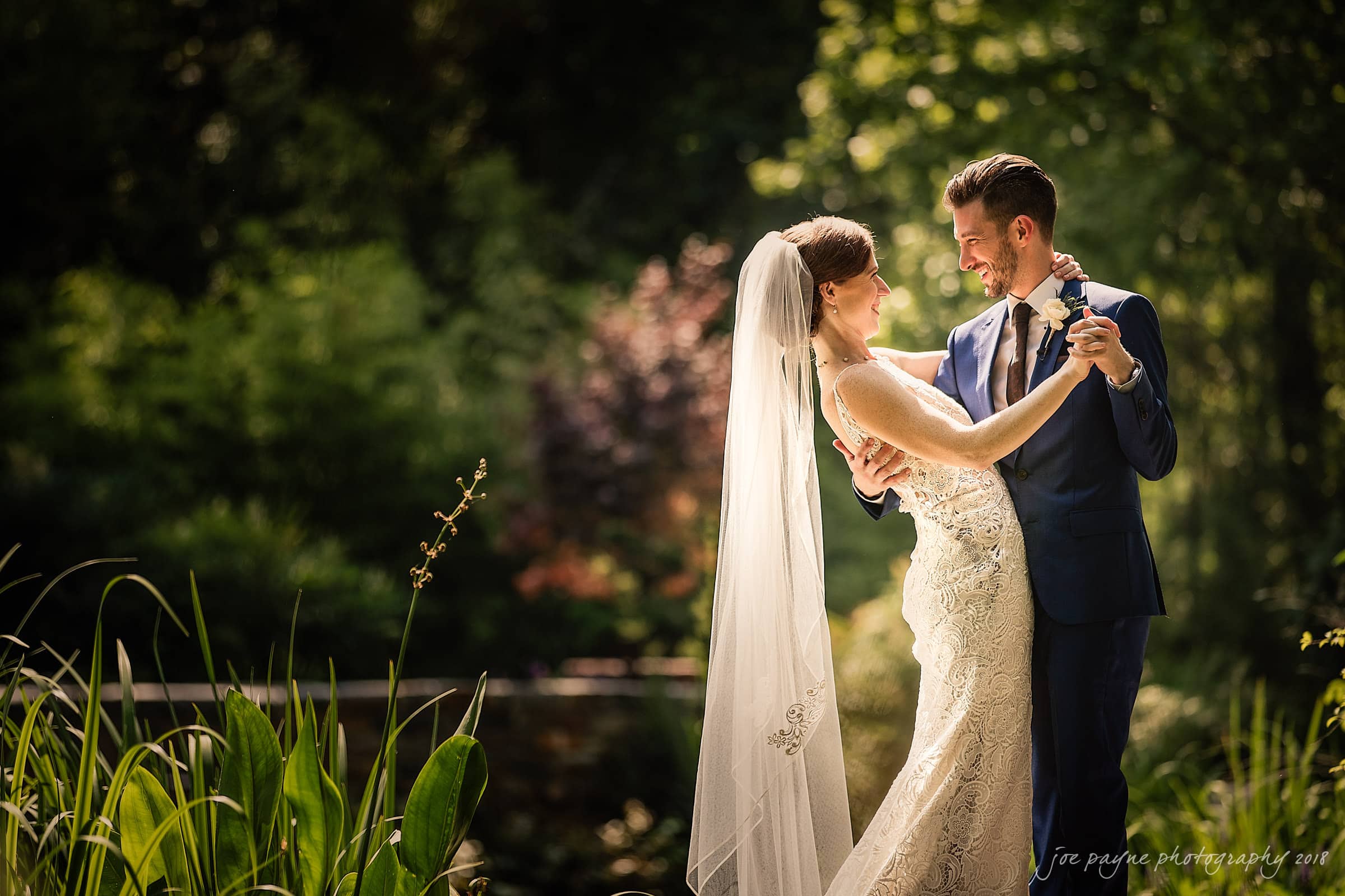 A bride in a white dress and veil and a groom in a blue suit dance outdoors surrounded by lush greenery. Sunlight filters through the trees, creating a serene and romantic atmosphere.