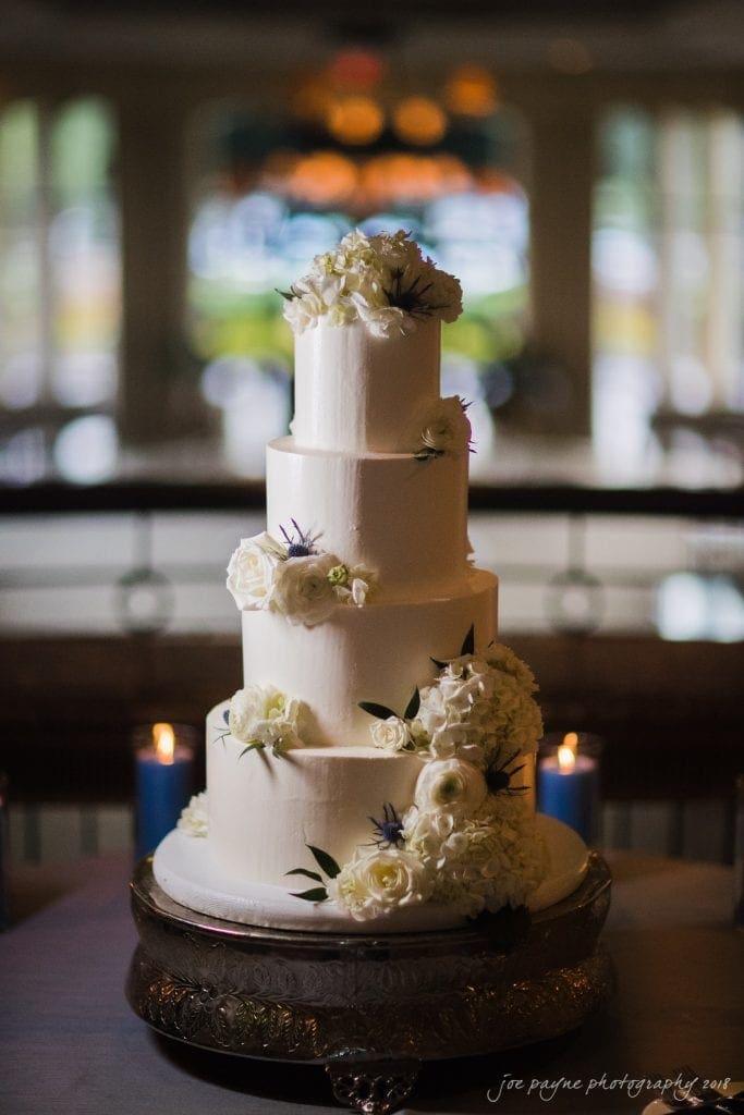 A four-tiered white wedding cake adorned with white flowers and greenery is displayed on an ornate silver stand. Three blue candles are lit in the background, adding a soft glow to the elegant setting.