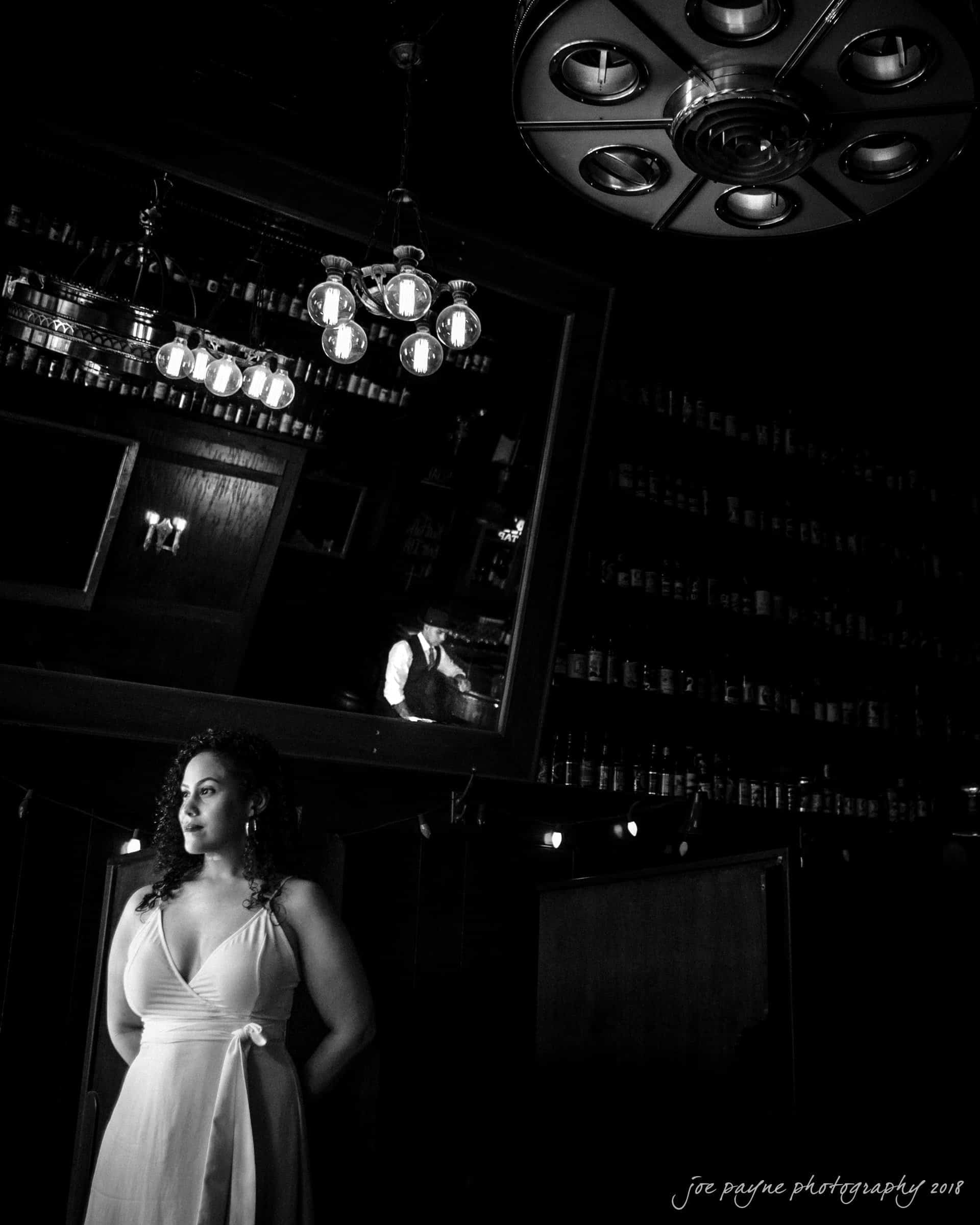 A woman in a white dress stands in a dimly lit room with vintage decor. A large mirror reflects shelves filled with bottles, and an ornate ceiling light fixture is visible. The scene exudes a classic, mysterious ambiance.