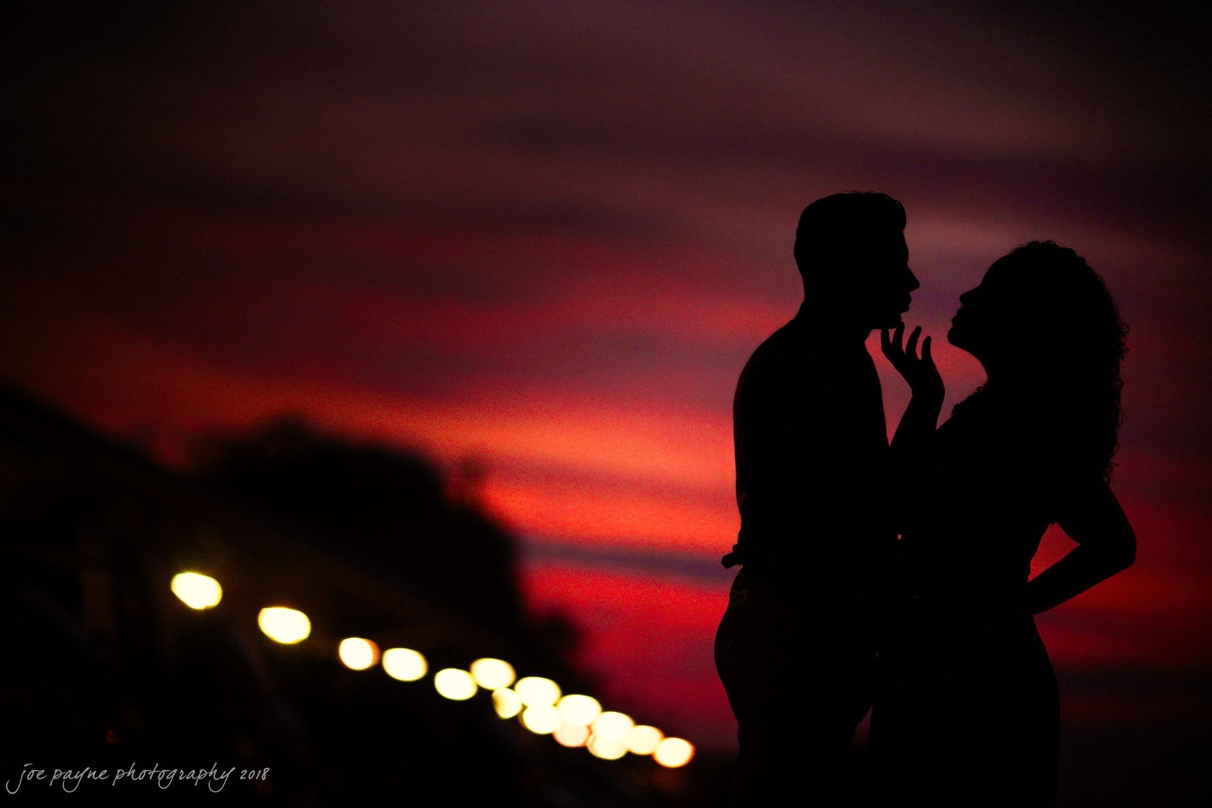 Silhouettes of a couple facing each other against a vibrant sunset sky. Soft glowing lights line a path in the background, adding a romantic atmosphere.