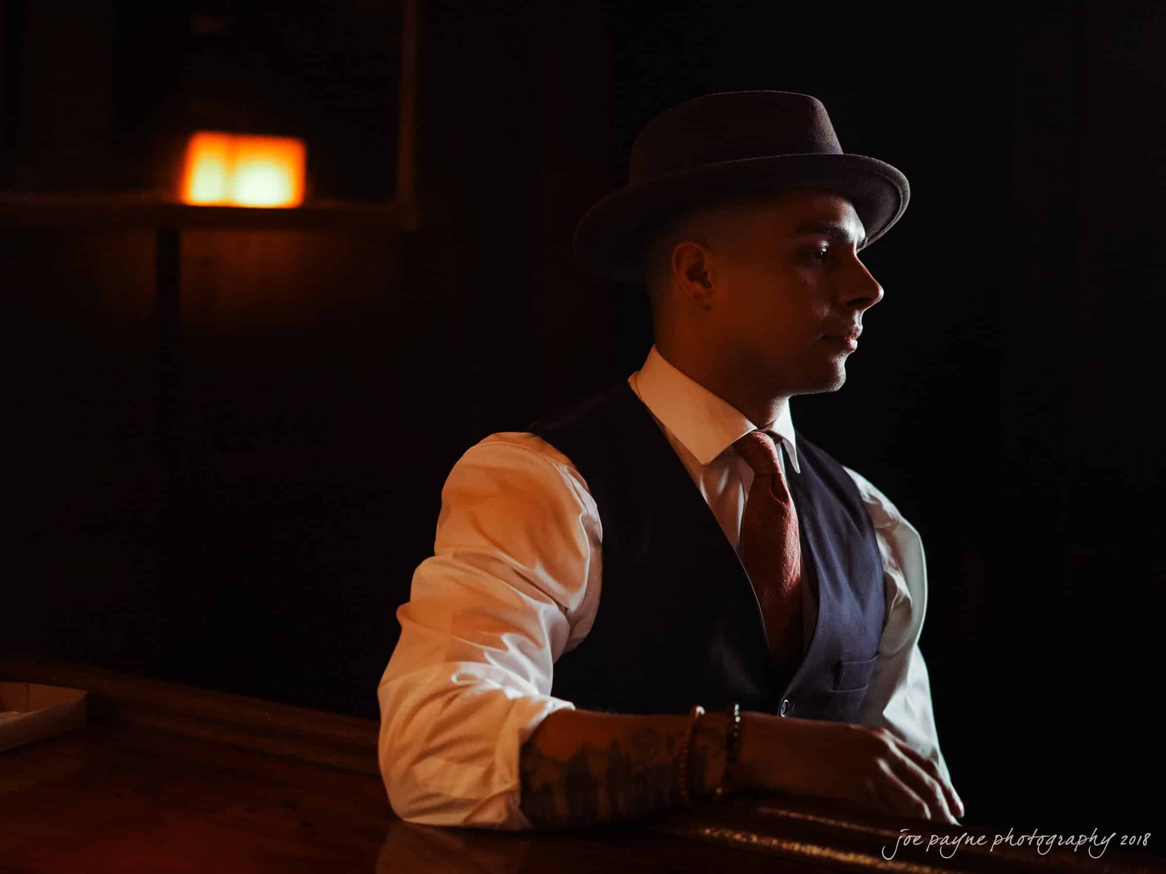 A person in a suit and fedora hat sits at a dimly lit bar, with a soft glow from a lamp in the background. They have tattoos on their arm and are looking to the side, with a focused expression.