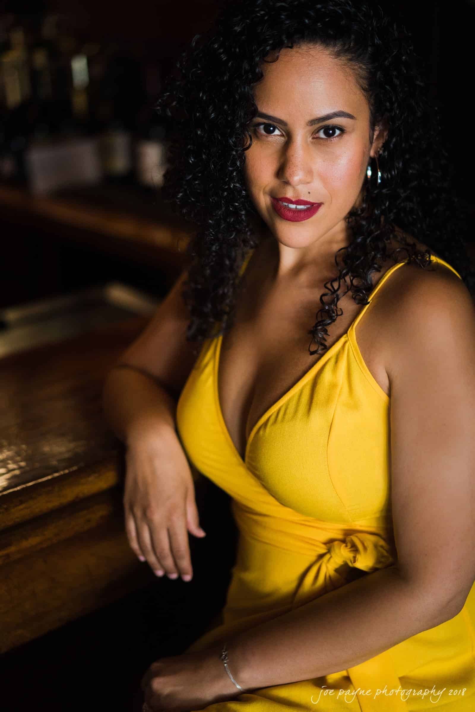 A woman with curly black hair wearing a yellow dress is leaning against a wooden surface. She has a confident expression and is looking directly at the camera. The background is softly blurred.