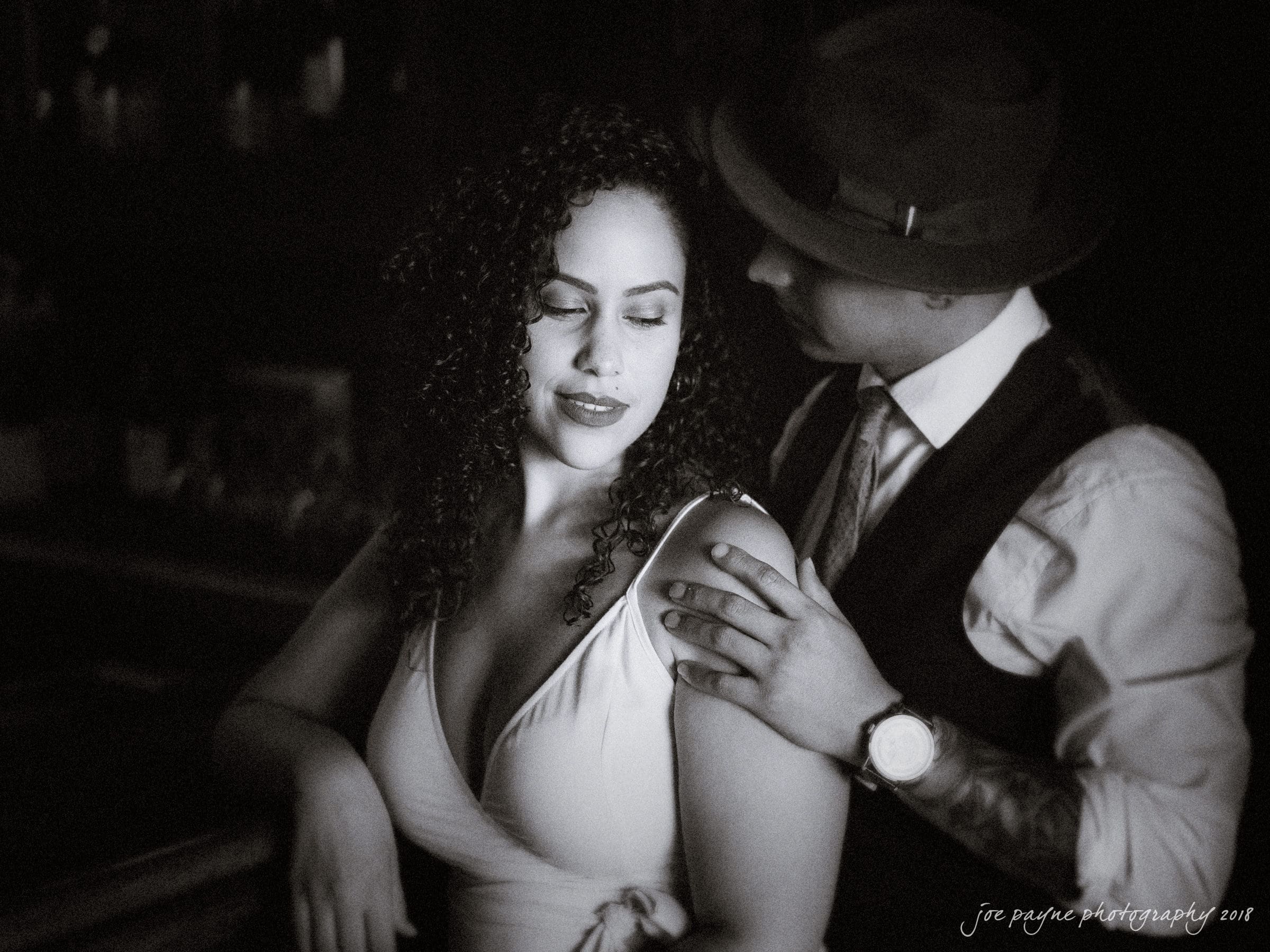A black and white photo of a man and woman in a close embrace. The woman, with curly hair and a sleeveless dress, gazes downwards. The man, wearing a hat and vest, looks at her affectionately while gently resting his hand on her shoulder.