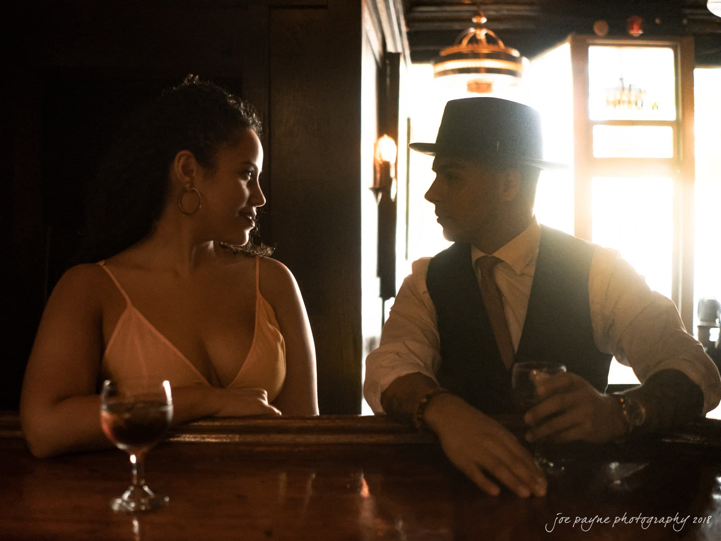 A woman and a man wearing a hat converse at a dimly lit bar, each holding a drink. Light from a window creates a silhouette effect. Hanging lights and wooden decor provide a warm ambiance.