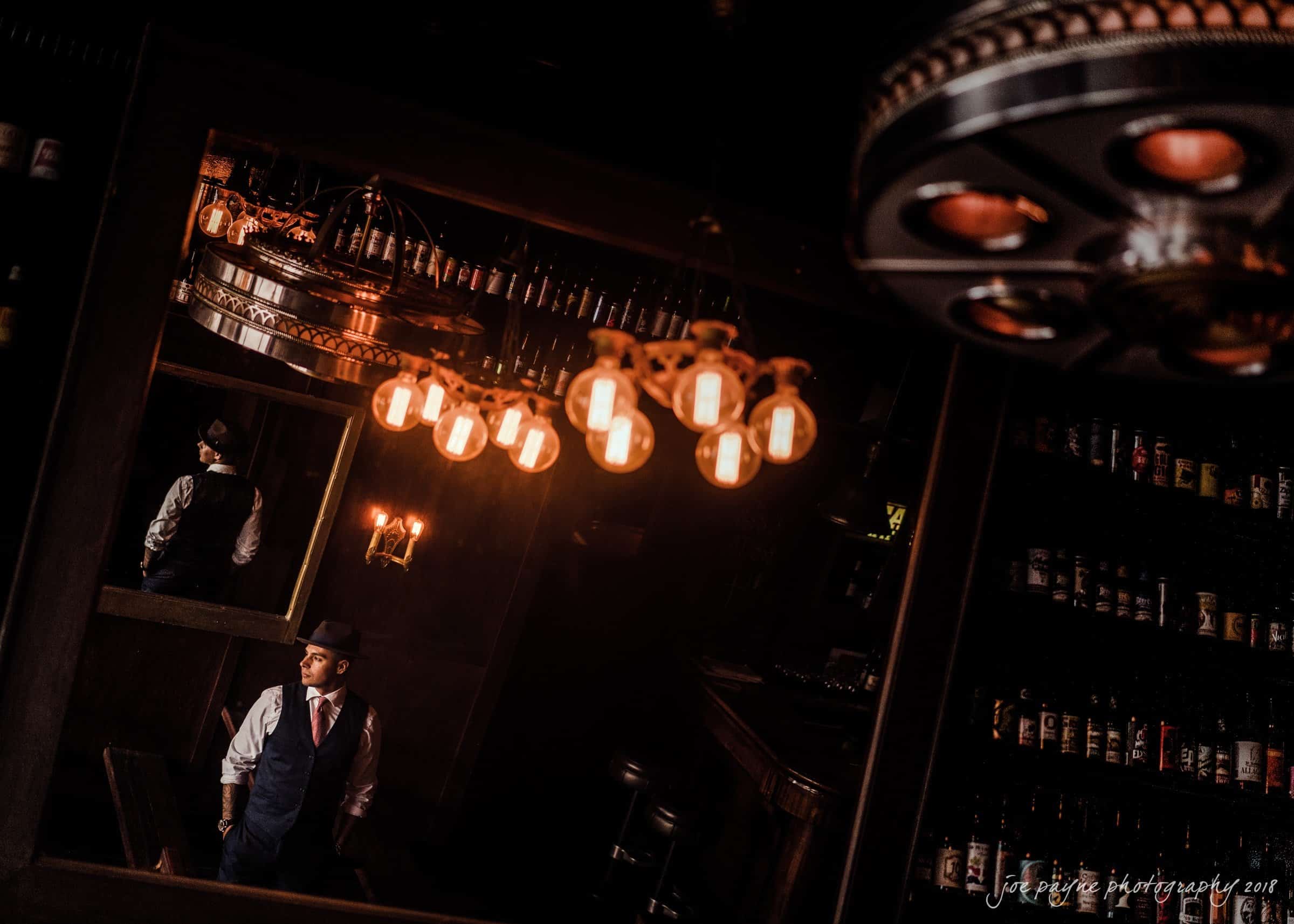 A man in a suit and fedora stands in a dimly lit bar, gazing towards the left. He is reflected in a mirror, surrounded by warm hanging lights and shelves of bottles. The setting has a vintage, atmospheric feel.