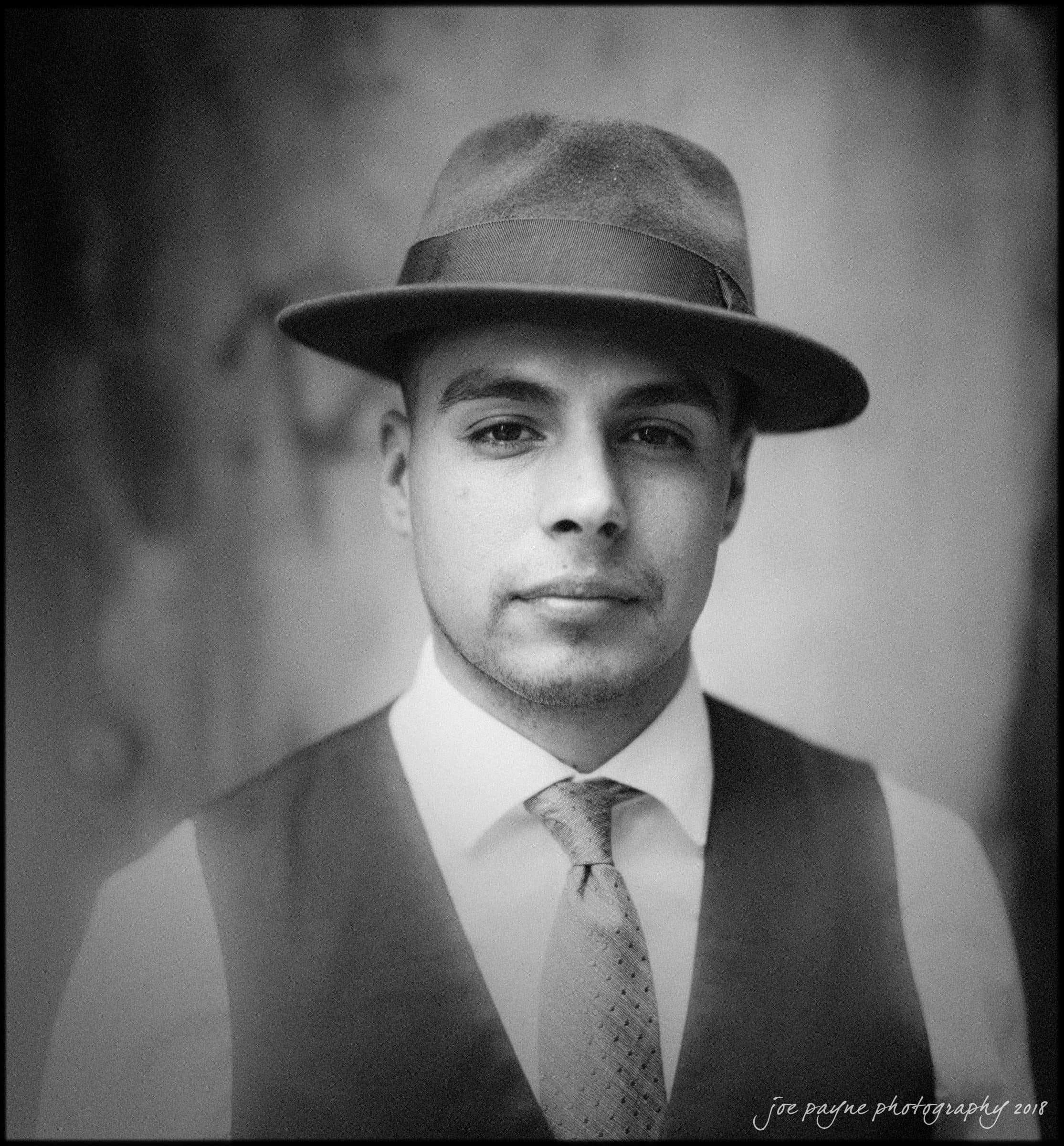 Black-and-white photo of a person wearing a hat and vest, with a neutral expression. The image is slightly soft-focused, giving it a vintage feel. The background is blurred.