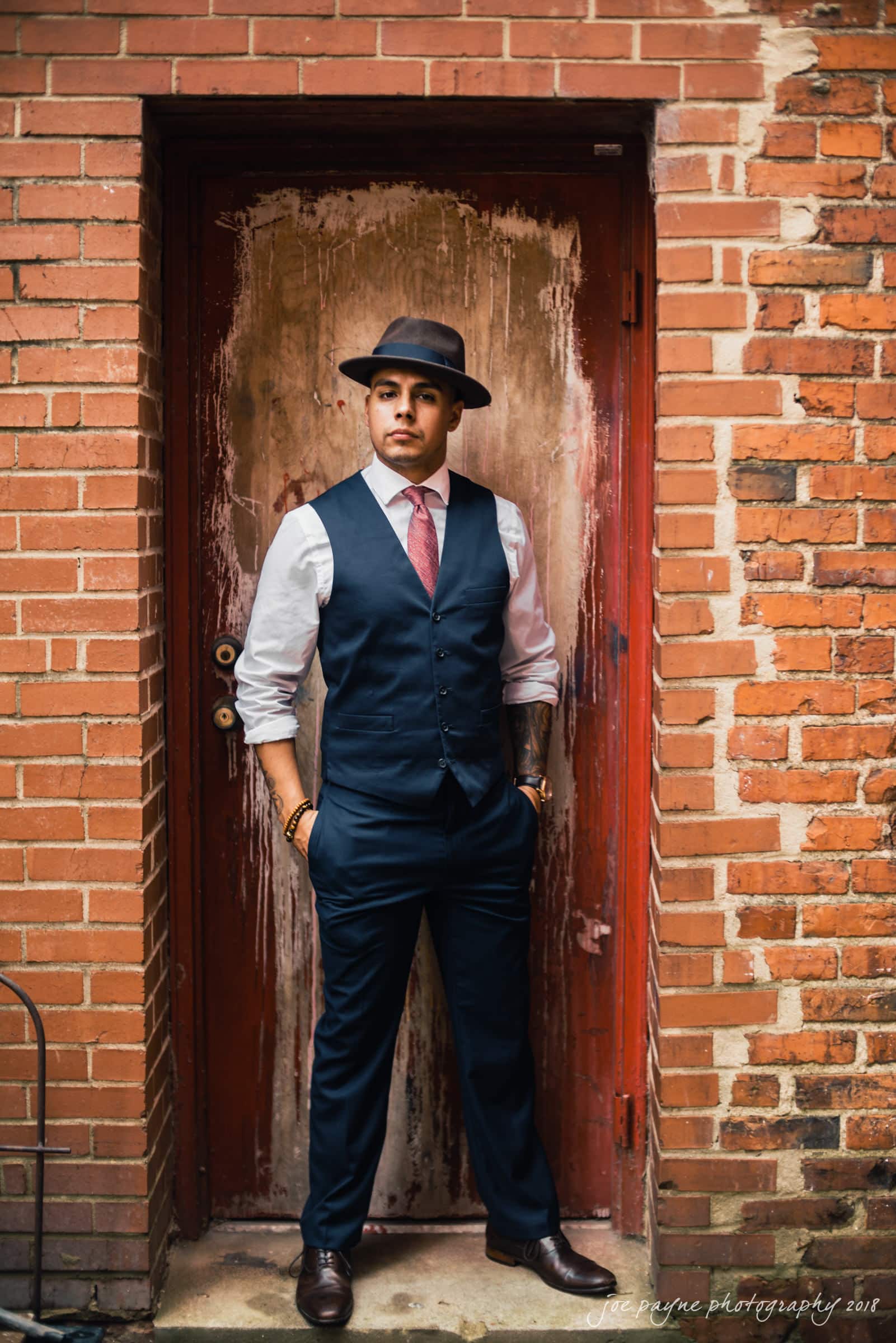 A man stands confidently in a narrow brick alleyway, wearing a fedora, a white shirt, a tie, and a dark vest with matching pants. His hands are in his pockets as he gazes forward. The backdrop is a distressed wooden door.