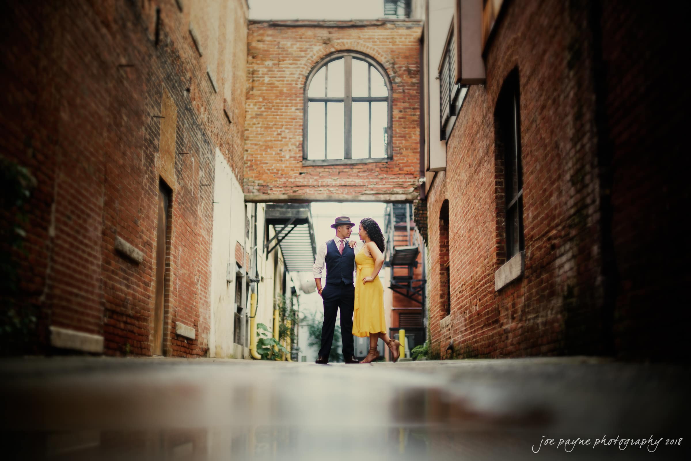 A couple stands in a narrow alleyway between brick buildings. The man is in a suit and hat, and the woman wears a yellow dress. They face each other, creating a romantic atmosphere under a large arched window above.