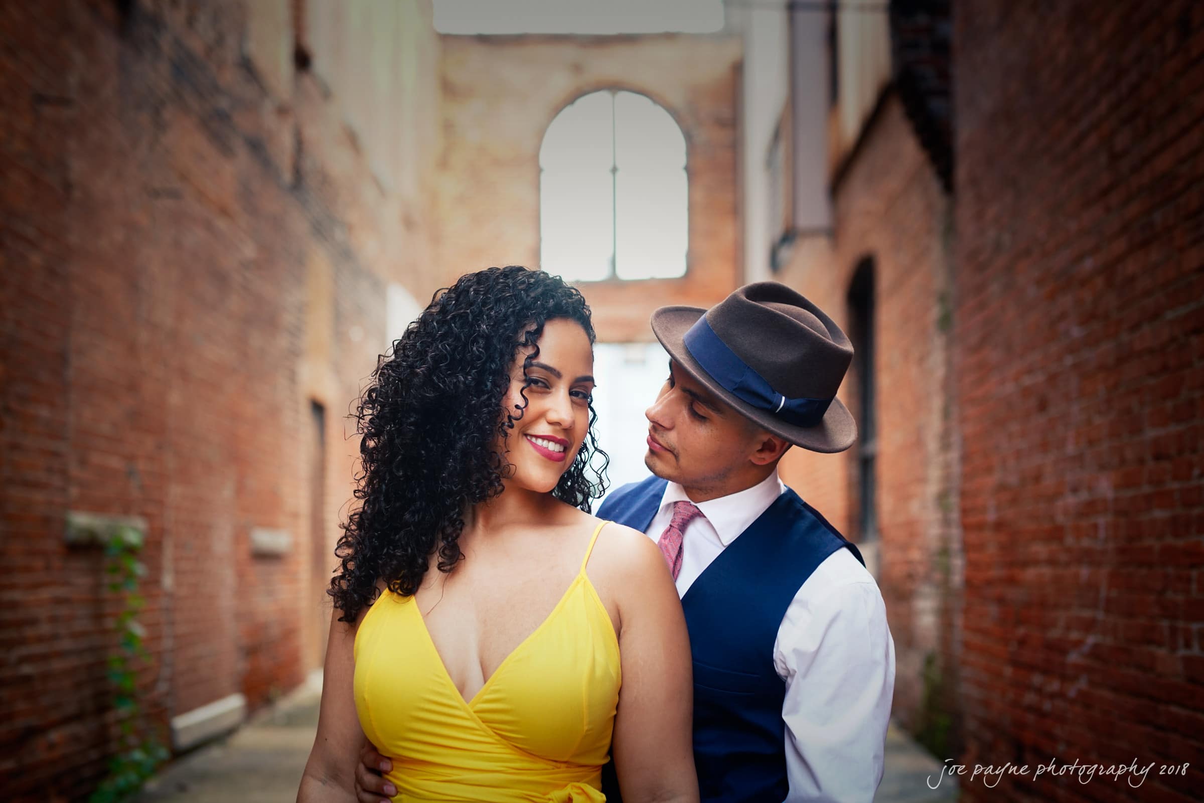 A couple stands in an alleyway between brick buildings. The woman, wearing a yellow dress, smiles. The man, in a hat and vest, gazes at her affectionately. An arched window with no glass is in the background.