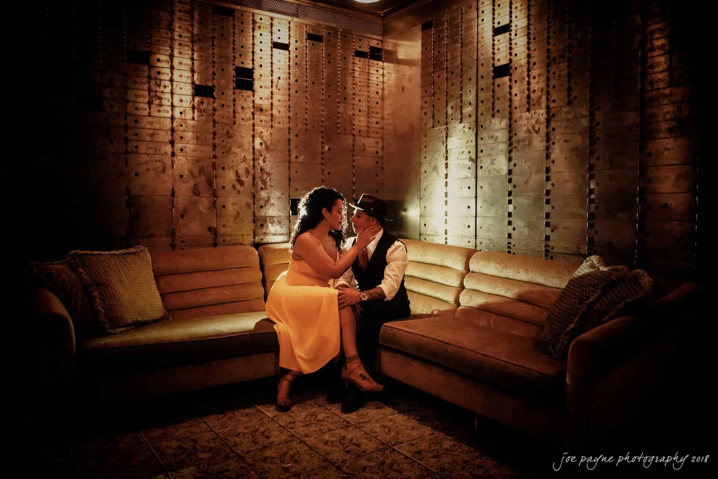 A couple sits on a luxurious couch in a dimly lit room. The woman in a yellow dress holds the mans chin while he wears a hat and vest. The background features a wall with a grid-like design, and the floor has a patterned tile.
