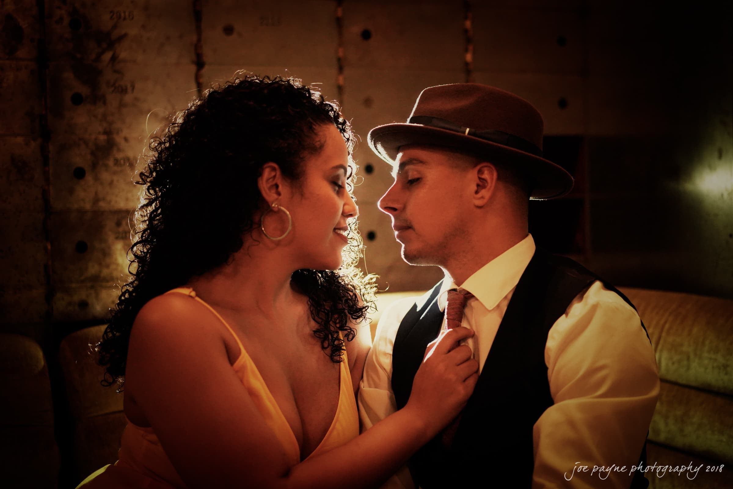 A couple in elegant attire shares an intimate moment in a dimly lit room. The woman wears a yellow dress and the man dons a hat, vest, and tie. They face each other closely, creating a warm and romantic atmosphere.