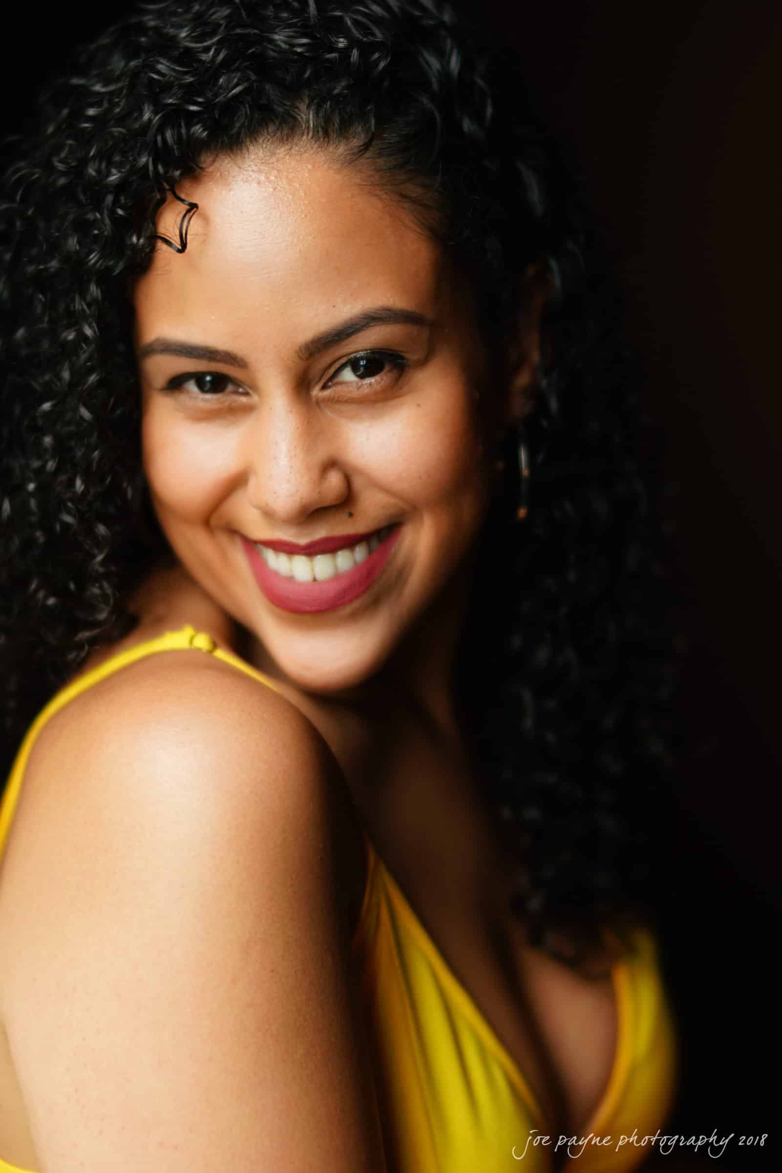 A person with curly hair smiles warmly while wearing a bright yellow top, set against a dark background.