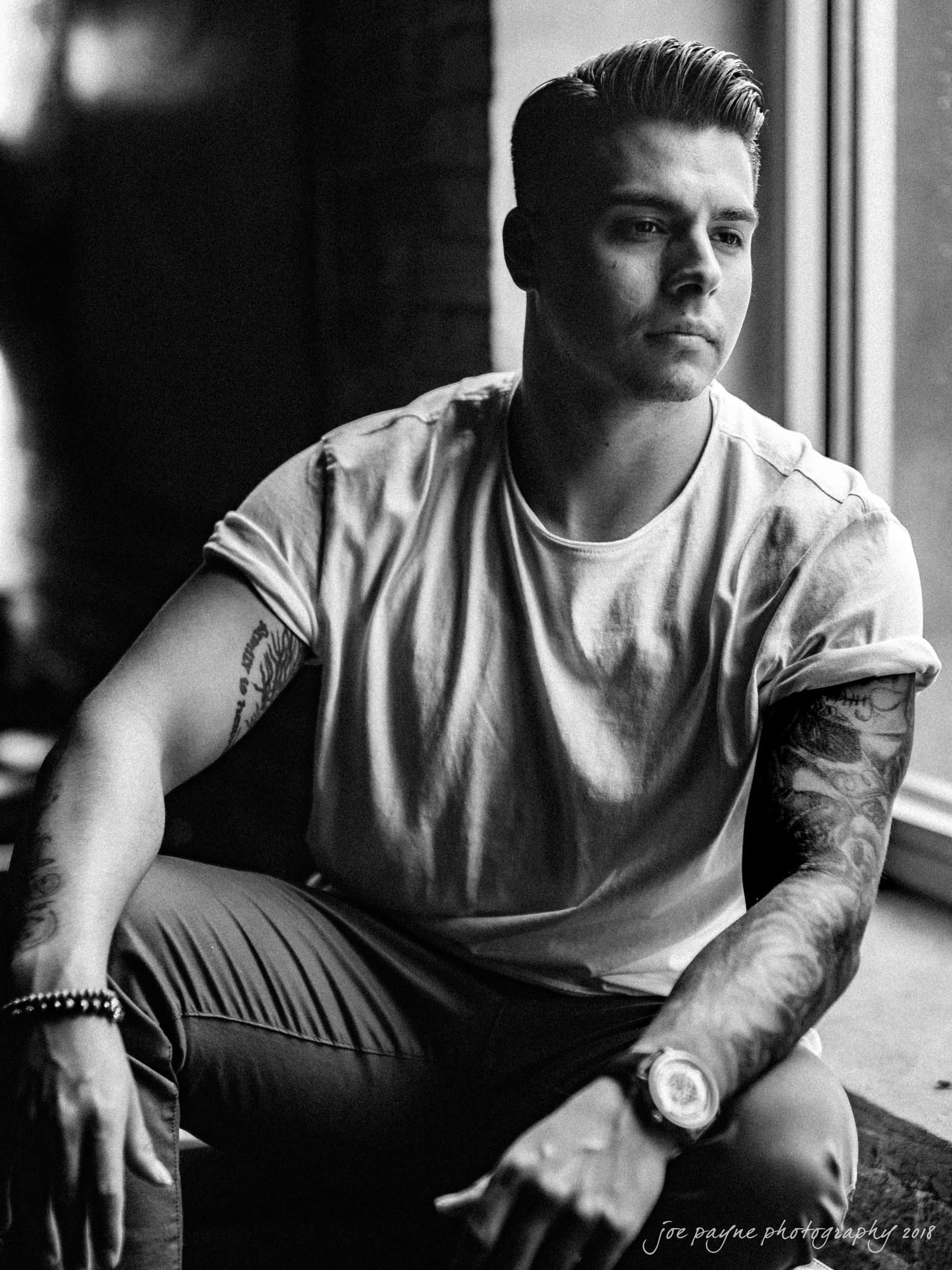Black and white photo of a man sitting by a window, wearing a T-shirt and pants. He has tattoos on both arms and is looking to the side, with light illuminating his face from the window.