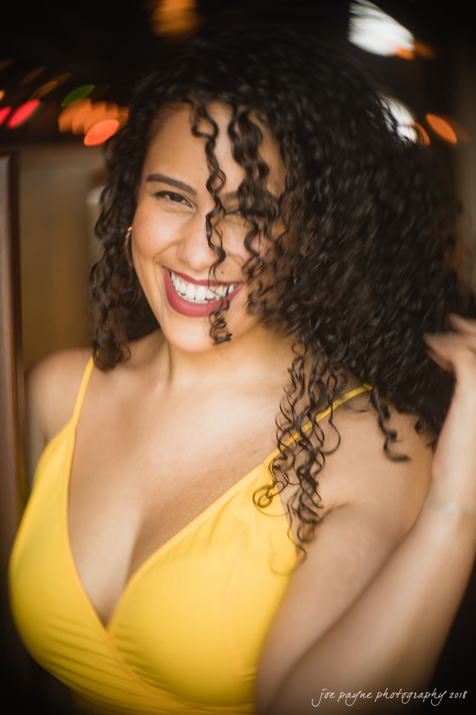 Woman with curly hair smiling brightly, wearing a yellow top. The background is blurred with colorful lights.