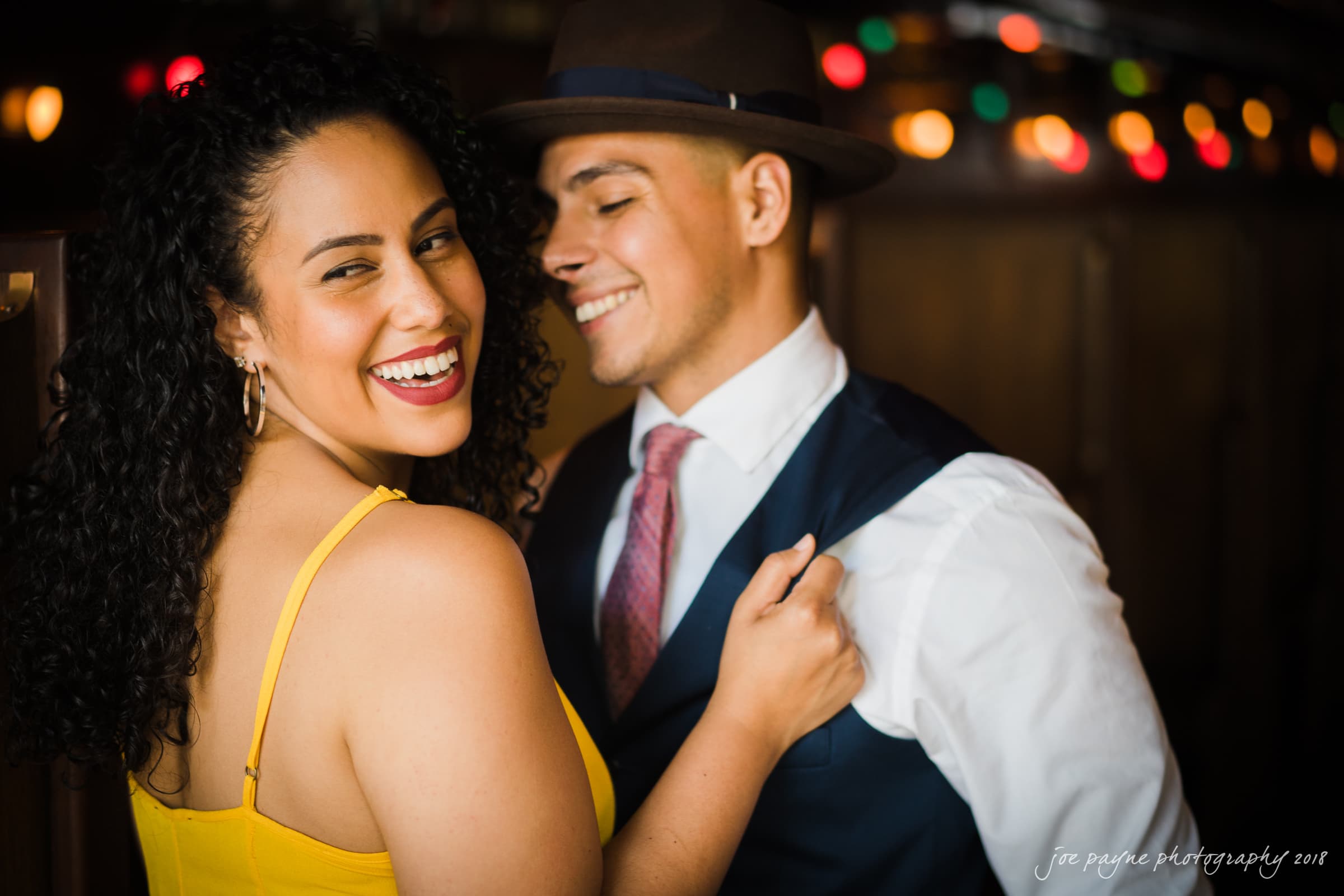 A woman in a yellow dress smiles at the camera while a man in a suit and hat stands beside her, smiling. They are in a warmly lit setting with colorful lights in the background.