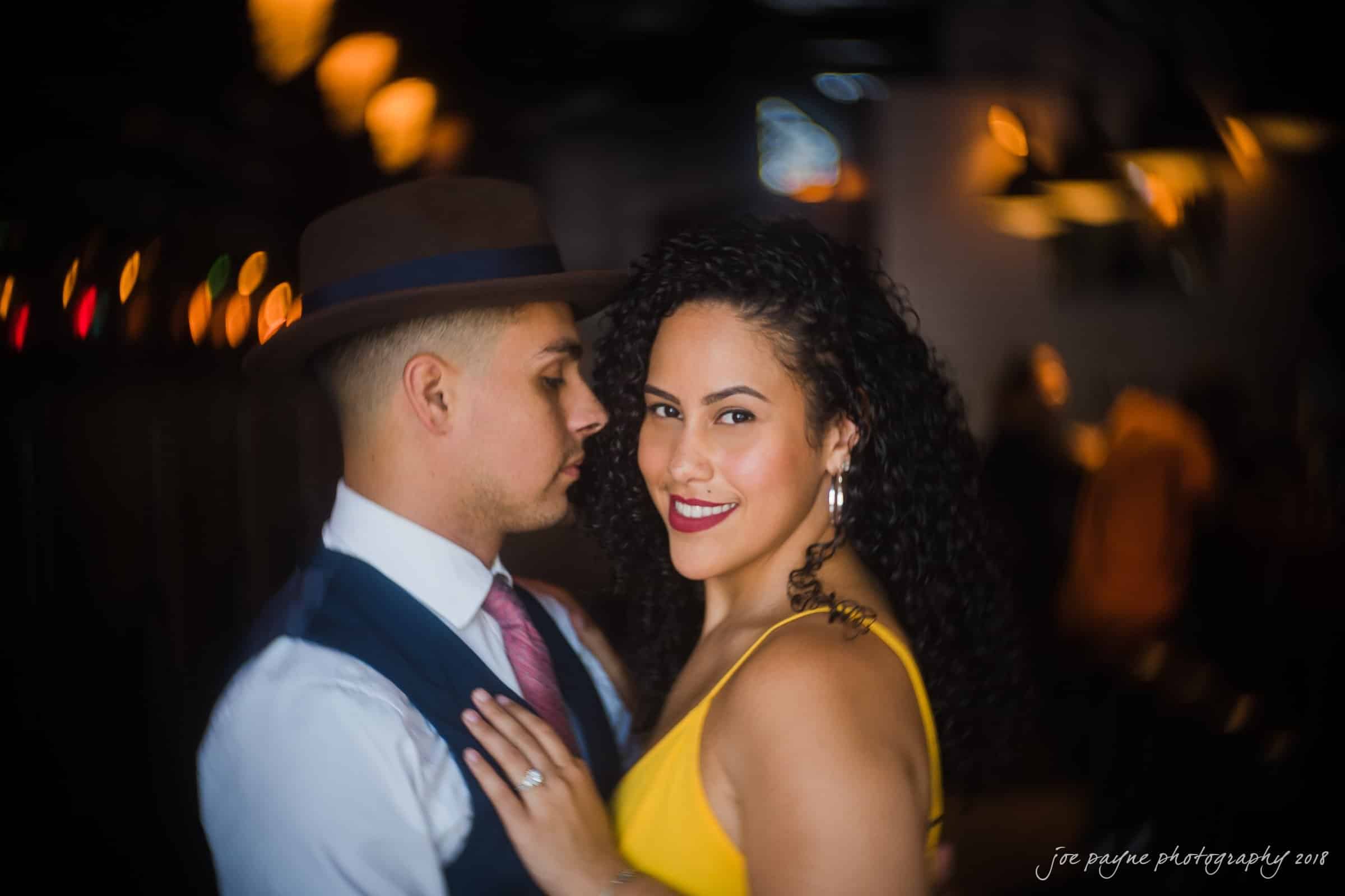 A couple poses closely in stylish attire, the man wearing a hat and a suit with a pink tie, and the woman in a yellow dress with curly hair, smiling. The background is softly blurred with warm, glowing lights.