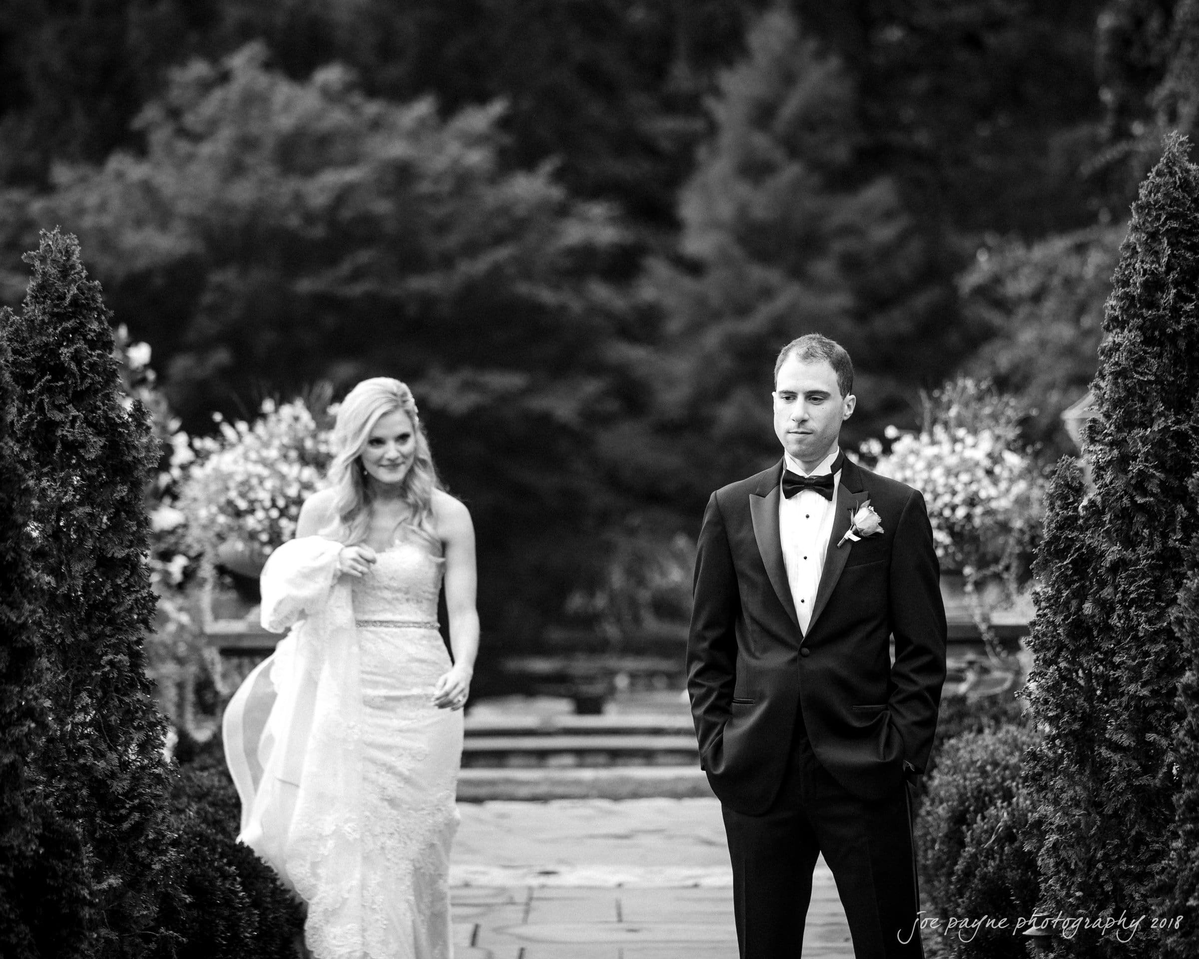 angle amphitheater at duke gardens wedding - katherine & andy A black and white photo of a bride and groom walking on a garden path. The bride is smiling and holding her dress, while the groom, dressed in a tuxedo, looks ahead. They are surrounded by lush greenery and flowers.