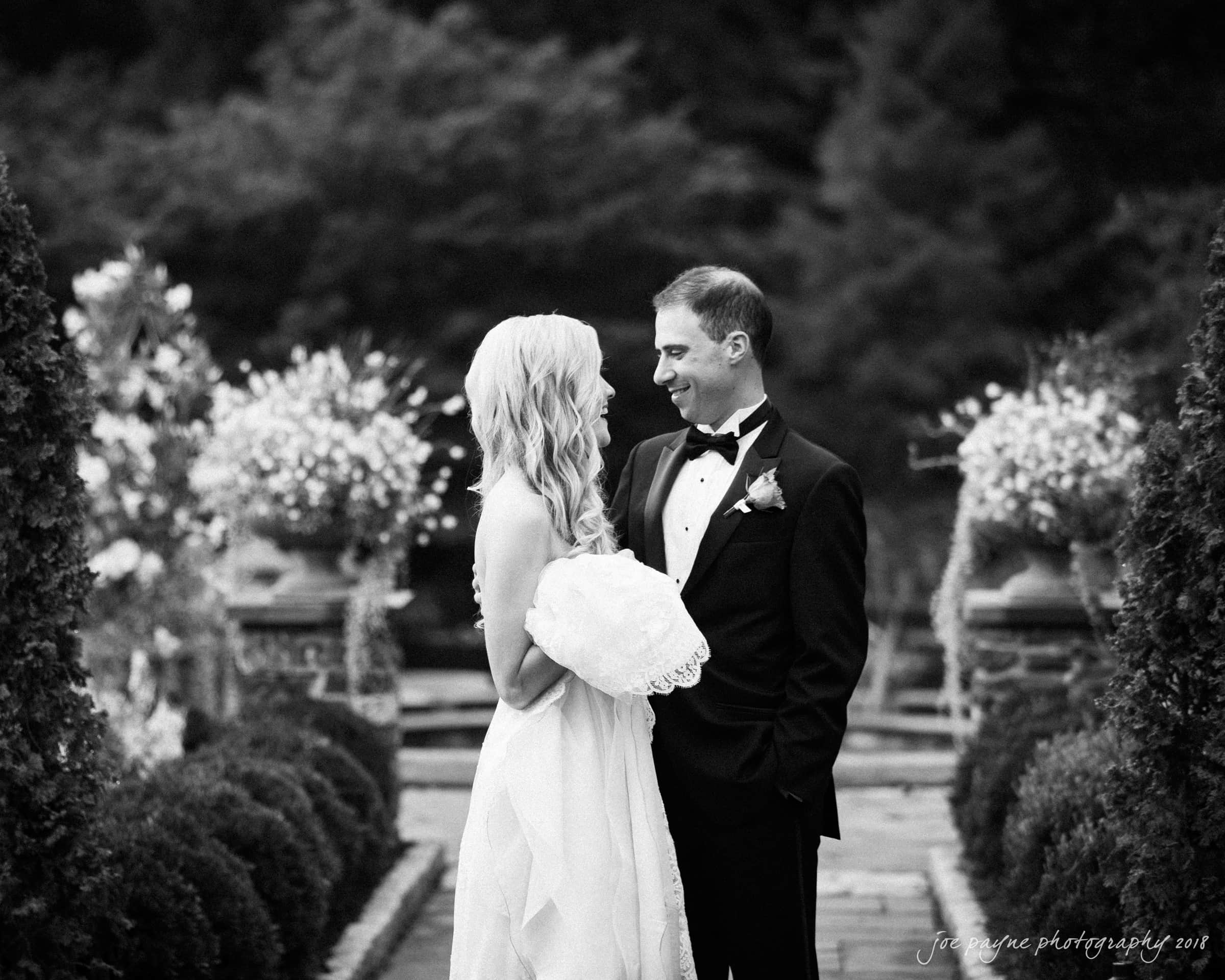 angle amphitheater at duke gardens wedding - katherine & andy A black and white photo of a bride and groom smiling at each other outdoors. The bride holds her dress, and the groom wears a tuxedo. They stand in a garden setting with decorative plants and blurred foliage in the background.