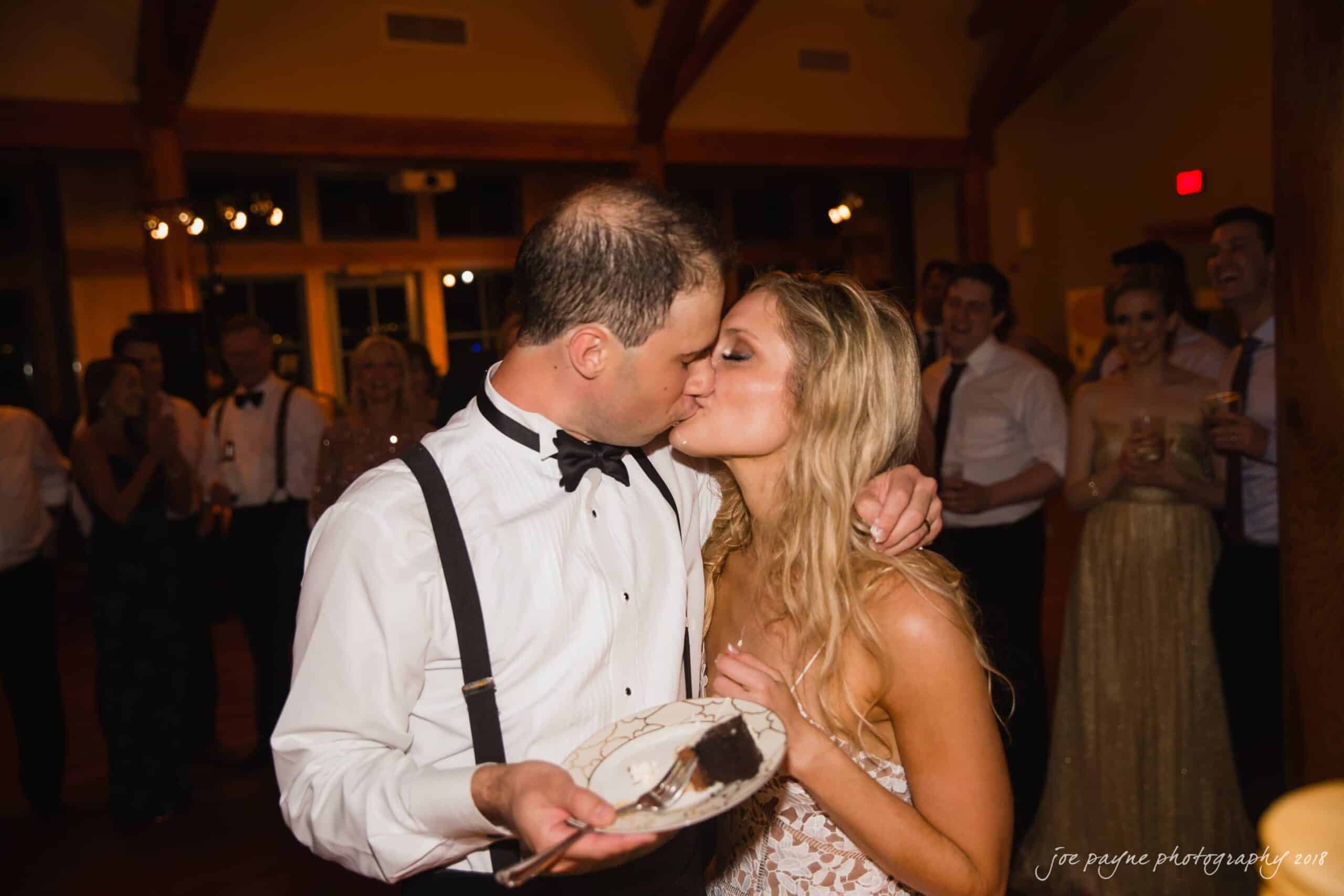 angle amphitheater at duke gardens wedding - katherine & andy A couple shares a kiss at a party, with the man holding a plate and fork with dessert. They are surrounded by dressed-up guests, visible in the background, who are smiling and enjoying the celebration.