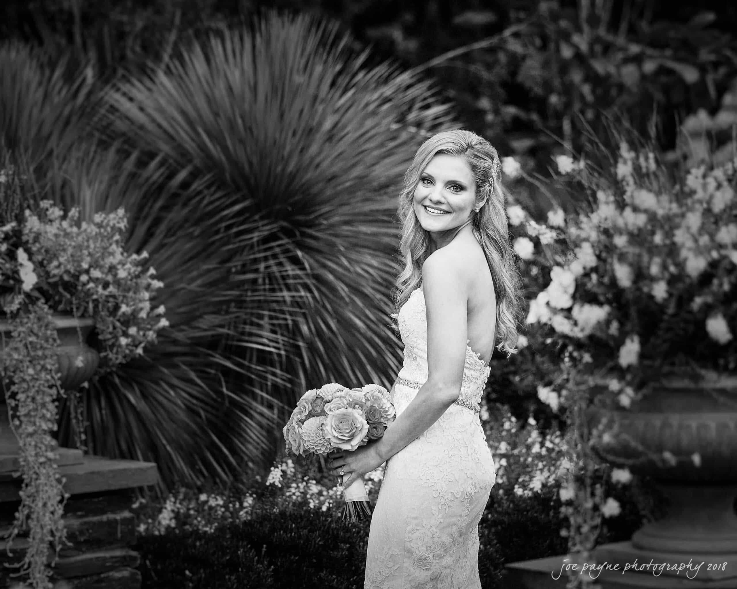 angle amphitheater at duke gardens wedding - katherine & andy A bride in a strapless lace gown holds a bouquet of roses and smiles while standing in a lush garden. The background features various plants and flowers, adding to the serene atmosphere. The image is in black and white.