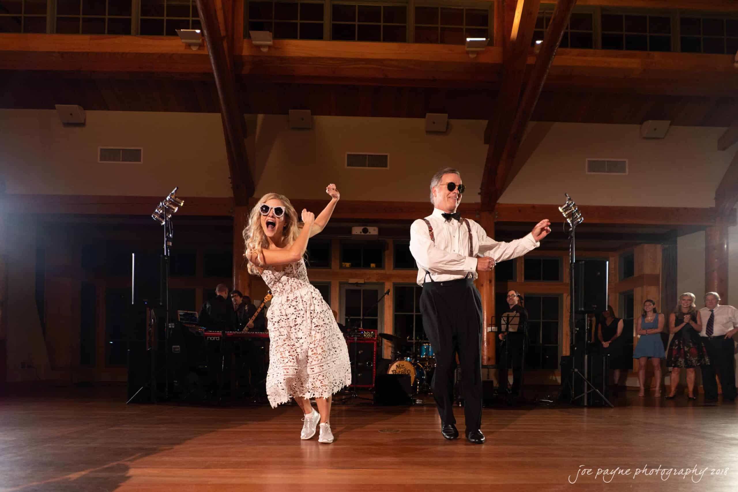 angle amphitheater at duke gardens wedding - katherine & andy A woman in a white dress and sunglasses dances energetically with a man in a tuxedo on a wooden dance floor. They appear joyful under bright stage lights with a band playing in the background.