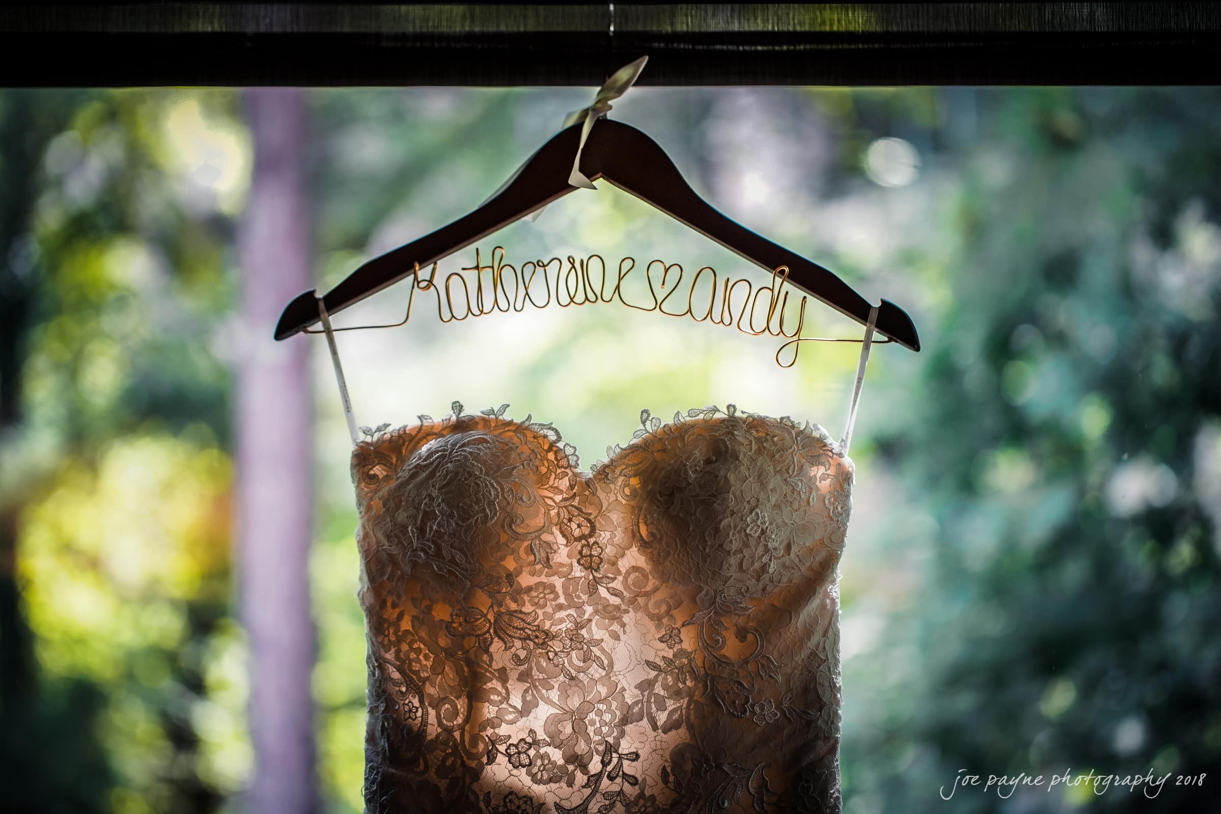 angle amphitheater at duke gardens wedding - katherine & andy A lace wedding dress hangs on a personalized hanger with the names Katherine and Andy in wire. The background is a blurred view of greenery through a window.