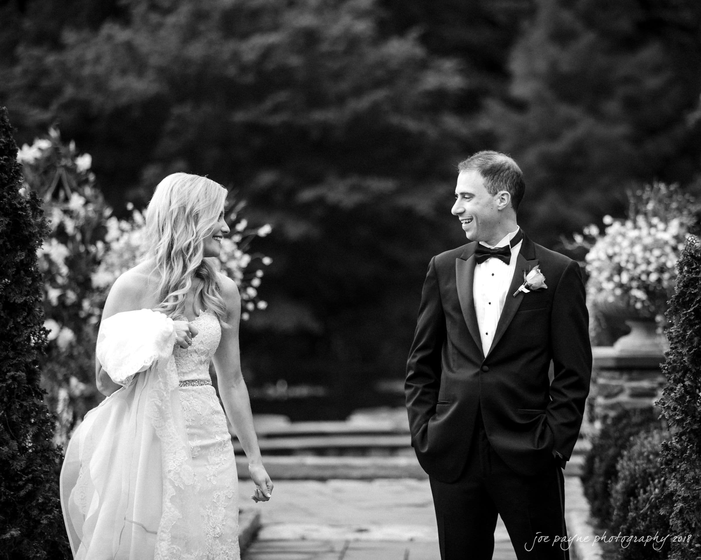 angle amphitheater at duke gardens wedding - katherine & andy A bride and groom share a happy moment outdoors. The bride is in a lace dress holding her train, while the groom wears a black tuxedo. They stand on a pathway surrounded by lush greenery and floral decorations, smiling at each other.