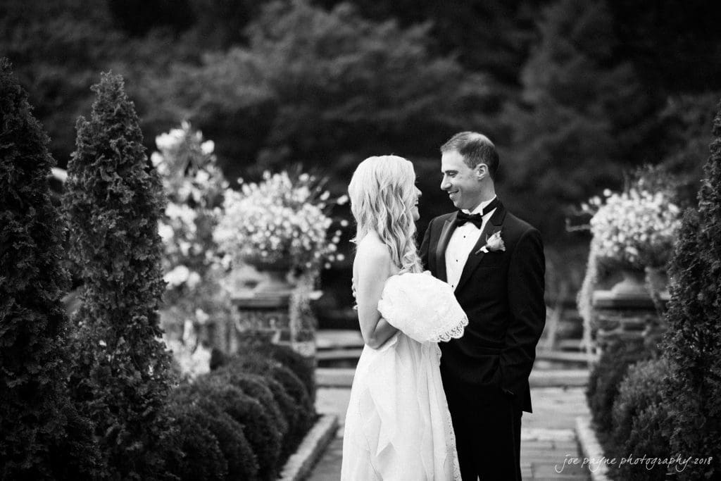 A black and white photo captures a couple in formal attire at Duke Gardens. The woman, adorned in a white dress, holds flowers while the man, in a suit, smiles fondly at her. They stand on a path lined with tall shrubs and ornate floral arrangements, encapsulating their wedding day's charm.