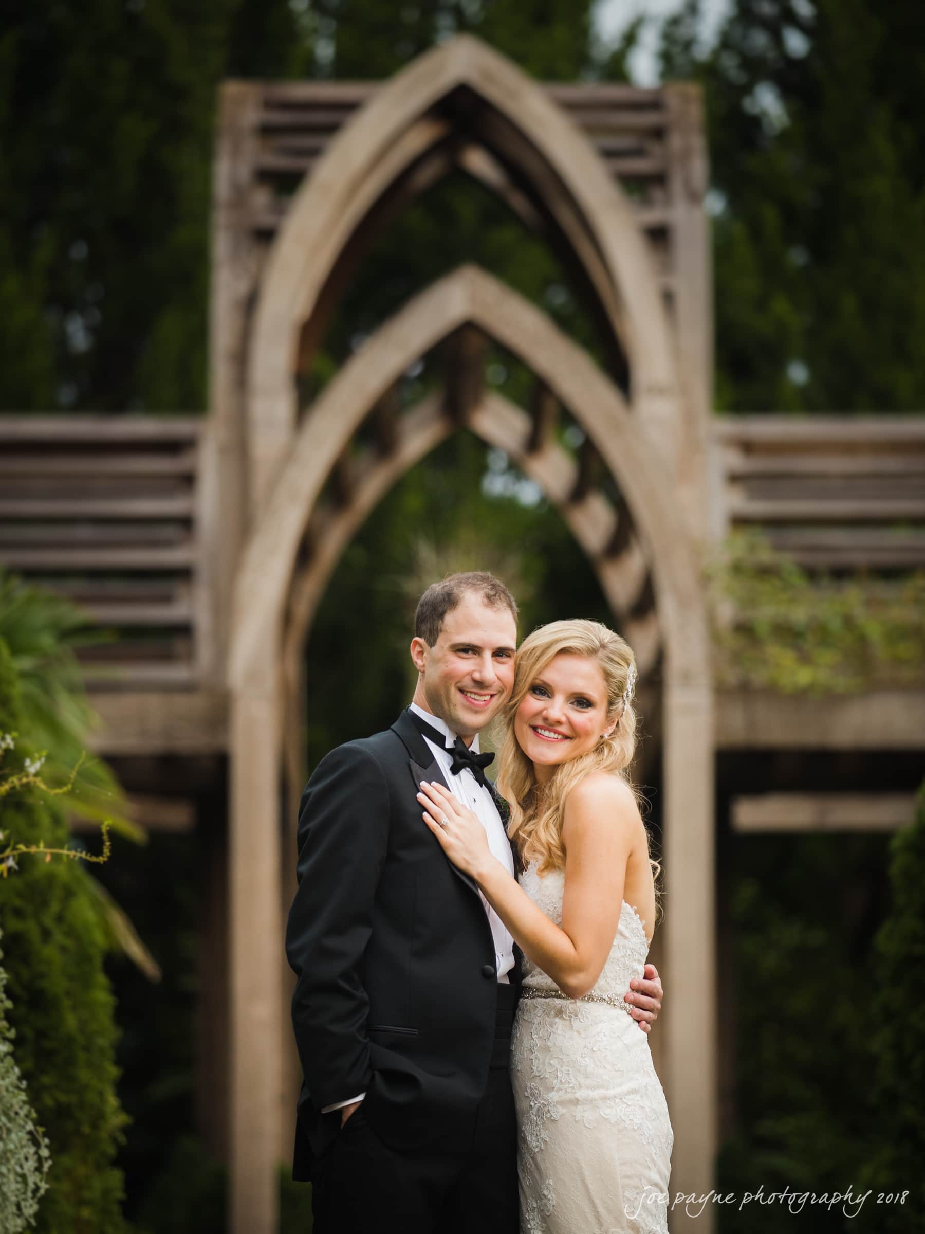 angle amphitheater at duke gardens wedding - katherine & andy A bride and groom smile while posing outdoors. The groom wears a black tuxedo, and the bride is in a white lace gown. They stand in front of an arched, wooden structure surrounded by greenery.