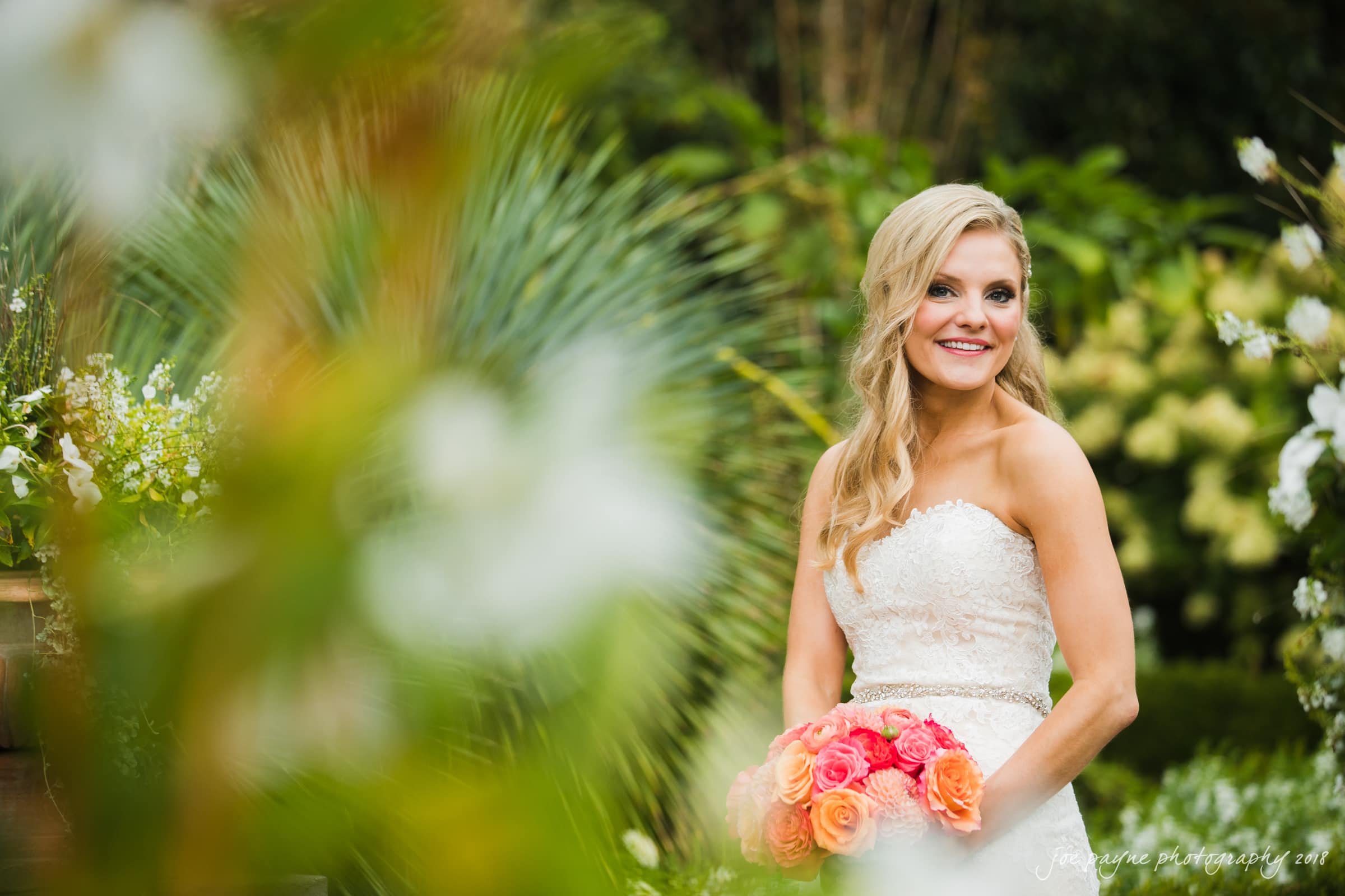 angle amphitheater at duke gardens wedding - katherine & andy A bride in a white strapless dress stands in a garden holding a bouquet of pink and orange roses. She has long blonde hair and smiles warmly, surrounded by lush greenery and blurred flowers in the foreground.