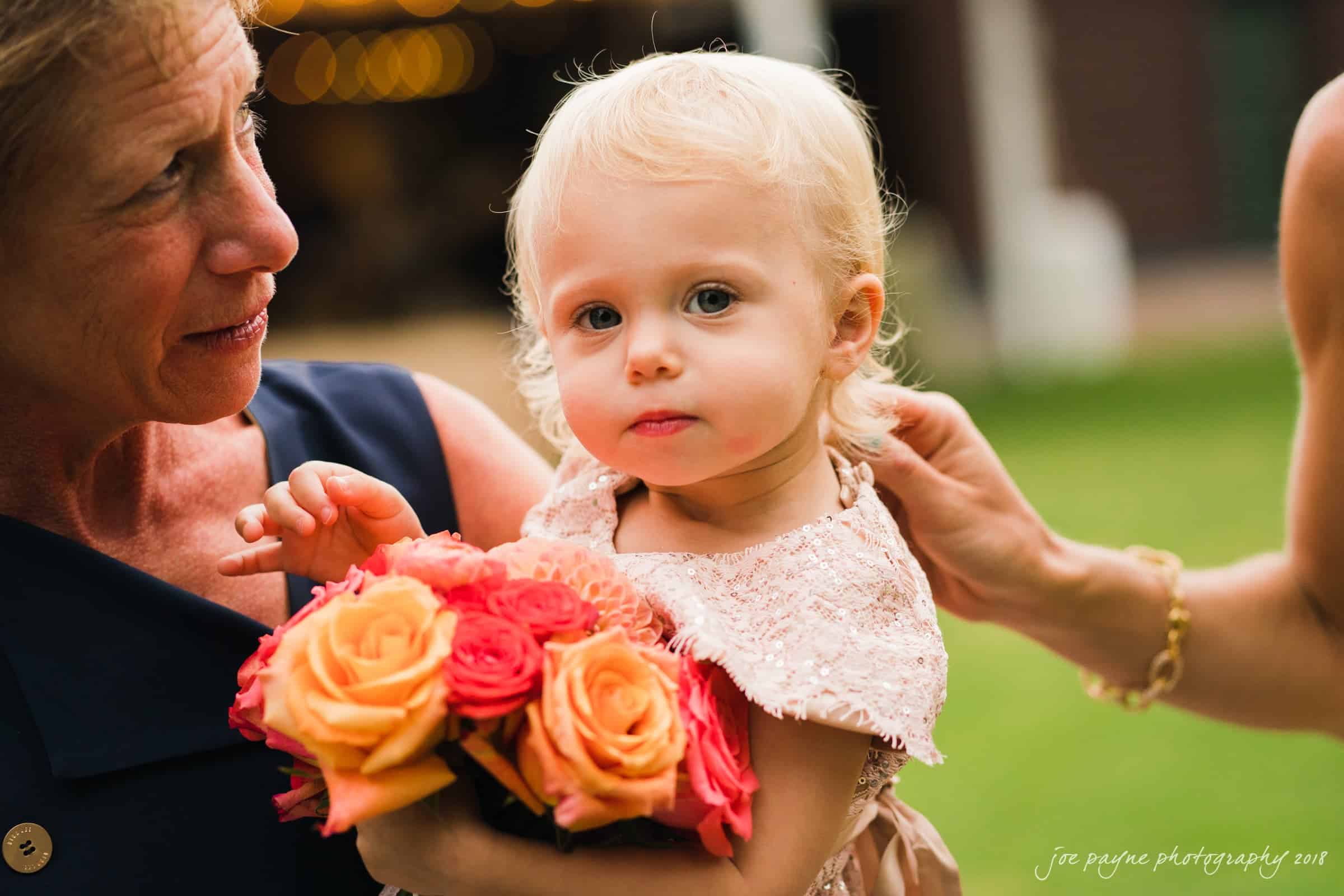 angle amphitheater at duke gardens wedding - katherine & andy A toddler with blonde hair holds a bouquet of orange and pink roses. An older woman, partially visible, interacts with the child, and another hand gently touches the toddlers back. The background is blurred with greenery.