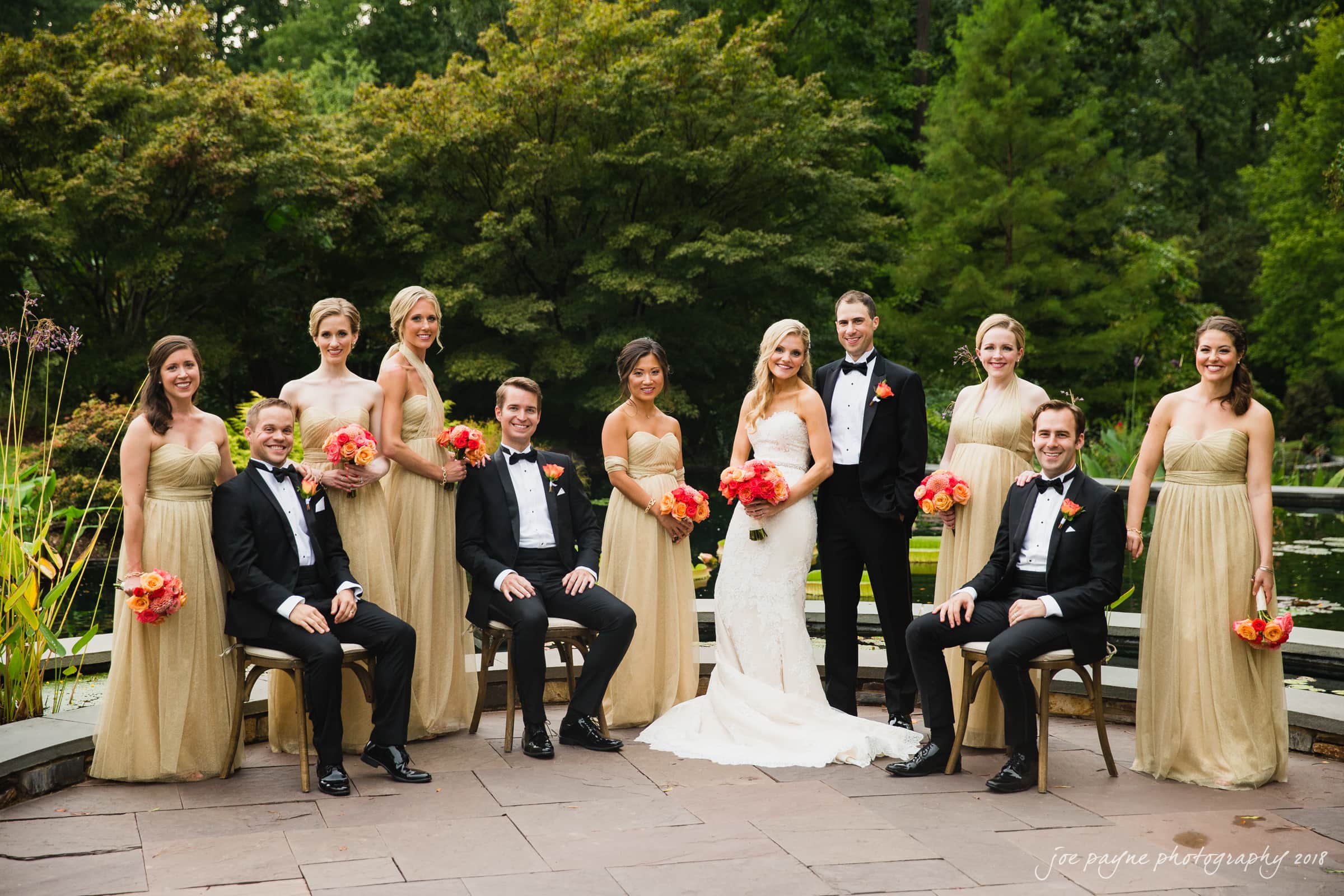 angle amphitheater at duke gardens wedding - katherine & andy A wedding party poses outdoors. The bride and groom stand in the center, surrounded by bridesmaids in gold dresses and groomsmen in black tuxedos. The bridesmaids hold orange and pink bouquets. A lush green garden serves as the backdrop.