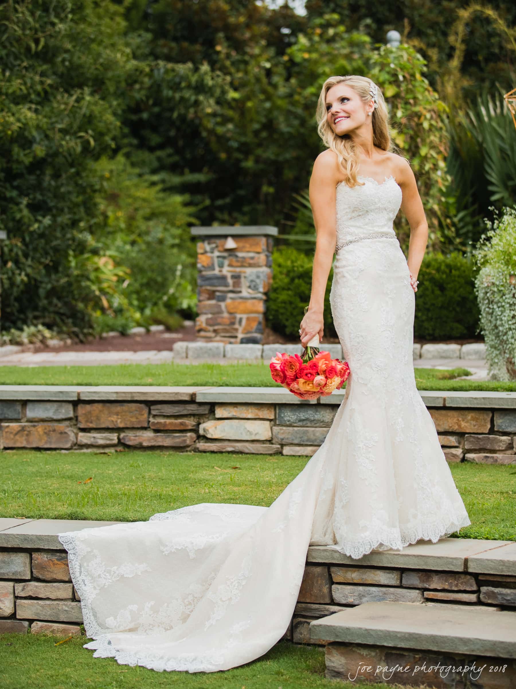 angle amphitheater at duke gardens wedding - katherine & andy A bride in a lace wedding gown stands outdoors on a stone pathway, holding a bouquet of orange and red flowers. She looks pleasantly to the side, with greenery and a stone wall in the background.