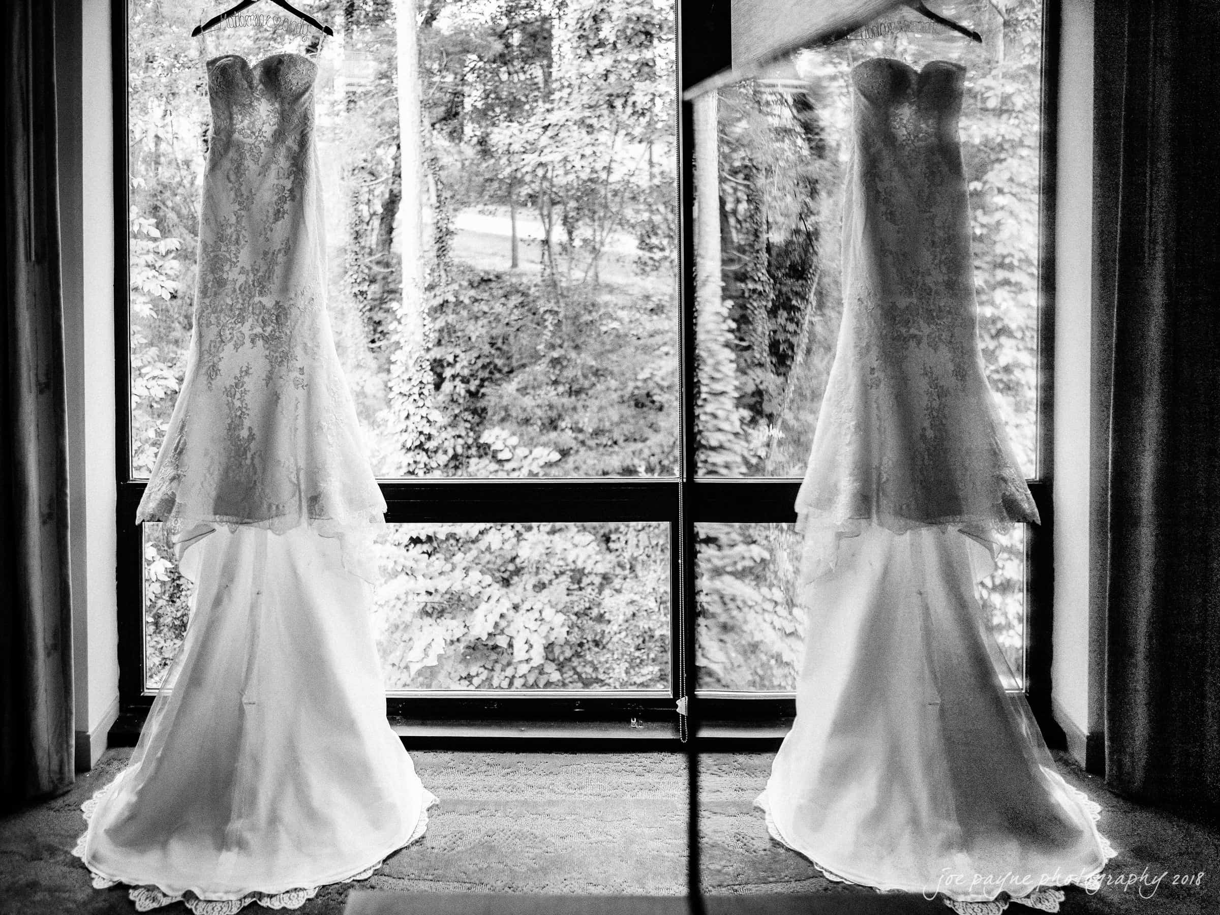 angle amphitheater at duke gardens wedding - katherine & andy Two elegant wedding dresses with lace detailing hang side by side in front of a large window. The window offers a view of a lush, wooded landscape outside. The image is in black and white, creating a timeless, classic feel.