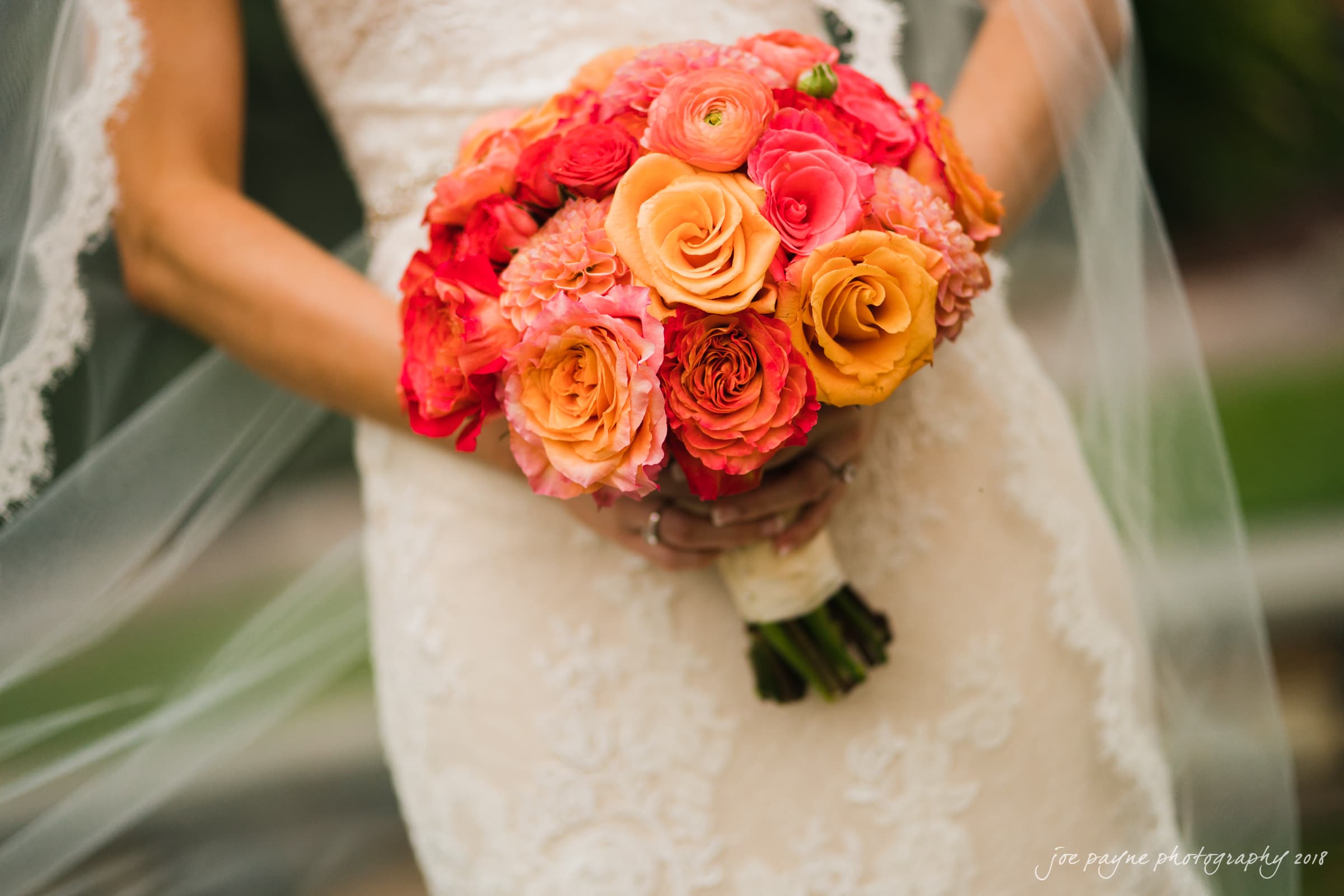 angle amphitheater at duke gardens wedding - katherine & andy A bride in a lace wedding dress holds a vibrant bouquet of orange and pink roses, with her veil draped gently over her arms. The background is softly blurred, highlighting the flowers.