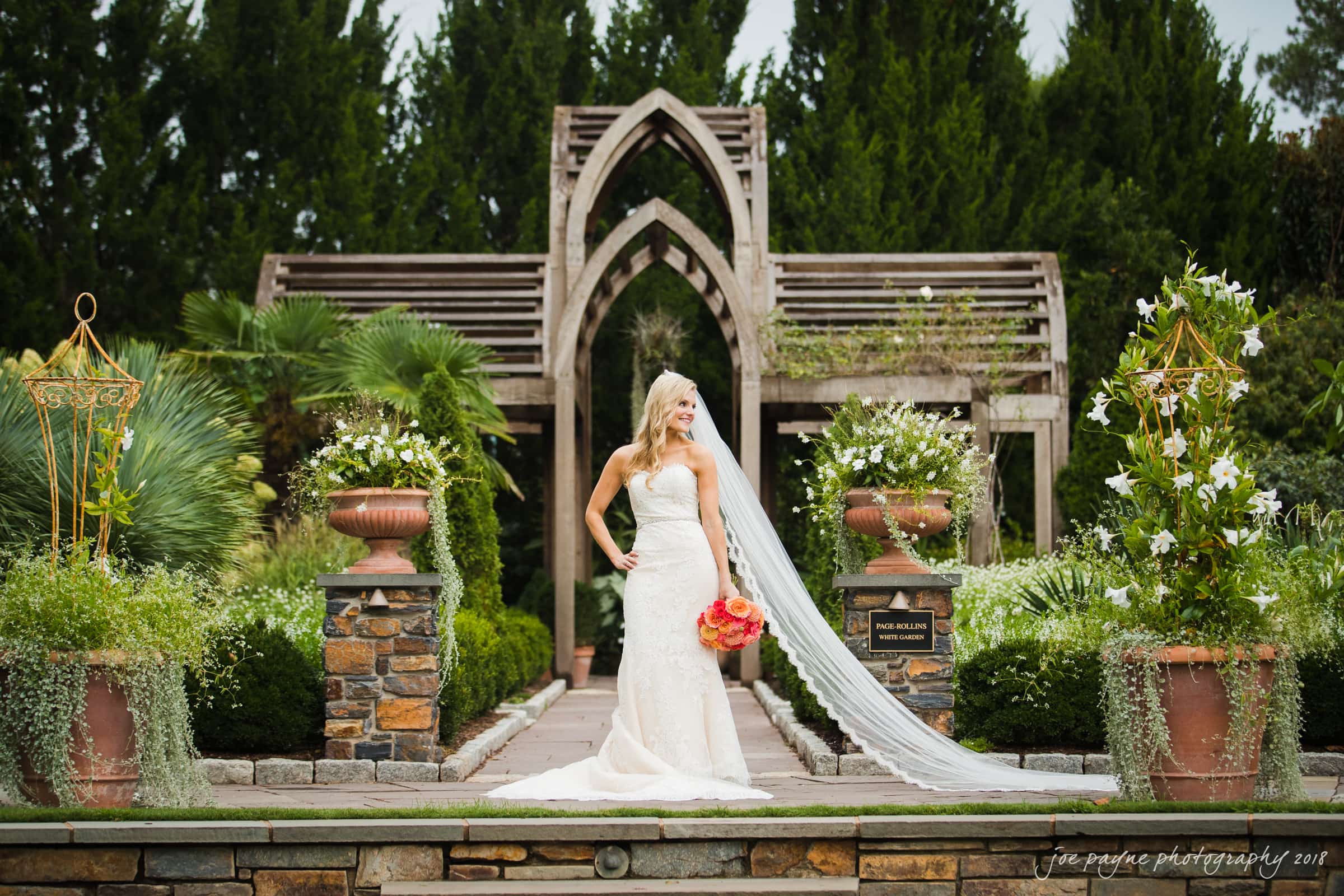 angle amphitheater at duke gardens wedding - katherine & andy A bride in a white gown and long veil stands in a lush garden, holding a vibrant bouquet of orange and pink flowers. Behind her is a wooden pergola surrounded by greenery and stone pillars.