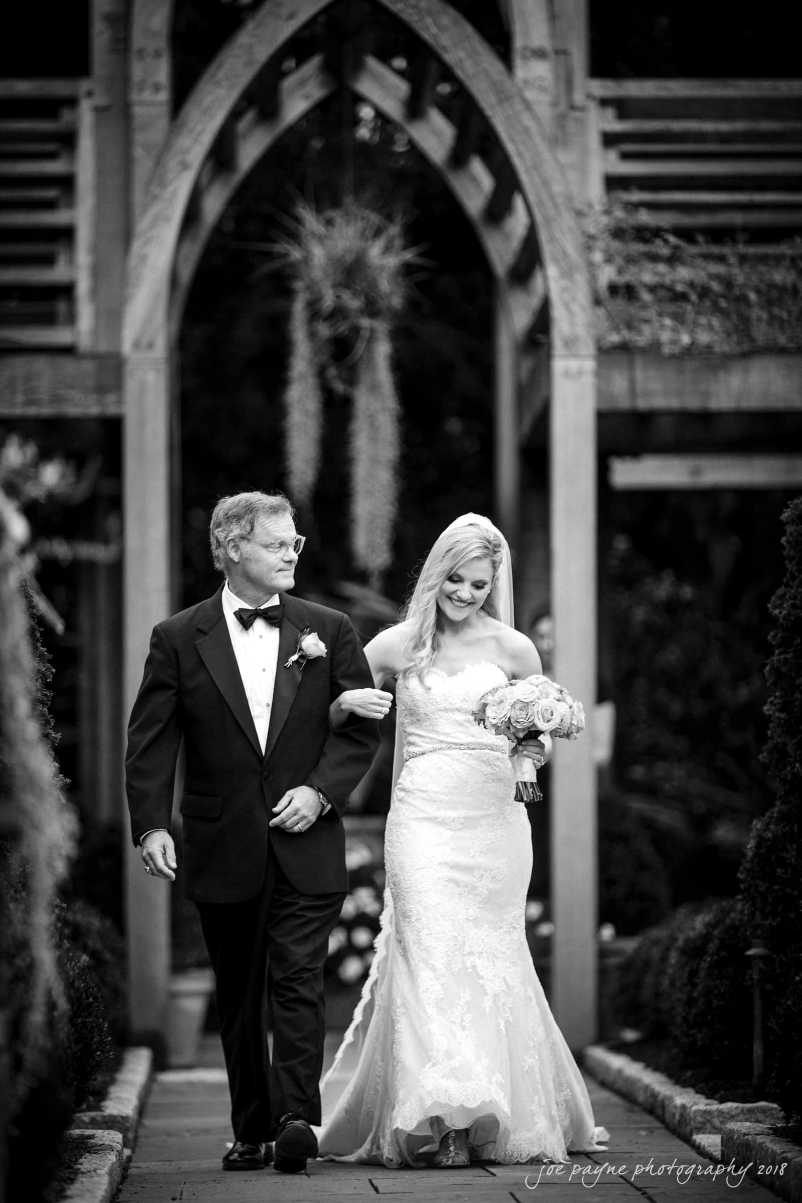 angle amphitheater at duke gardens wedding - katherine & andy A bride in a strapless gown holds a bouquet and walks arm-in-arm with an older man in a tuxedo. They are outdoors under a wooden archway in a garden setting. The image is in black and white.
